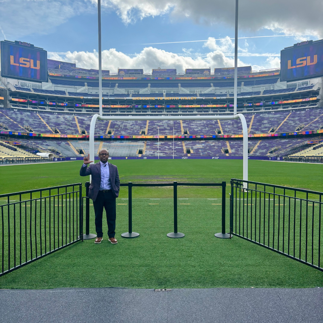 This is Dr. Dexter Shorter in a suit standing on a field near a goalpost in an empty football stadium with purple seats, digital screens displaying 'LSU' and a message 'Welcome to Death Valley' on a partly cloudy sky.