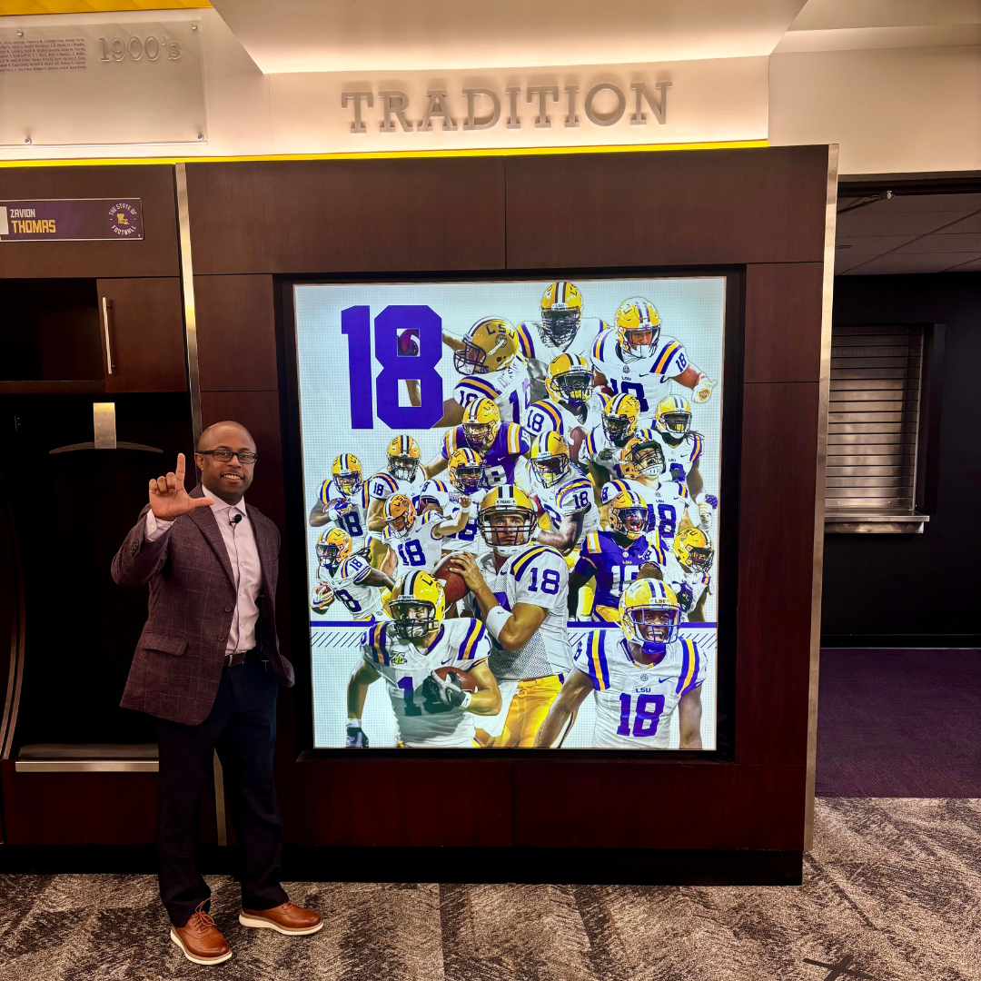 This is Dr. Dexter Shorter in a business suit posing with an "L" hand gesture next to a digital display of multiple football players in LSU uniforms. The display features the number 18 prominently.