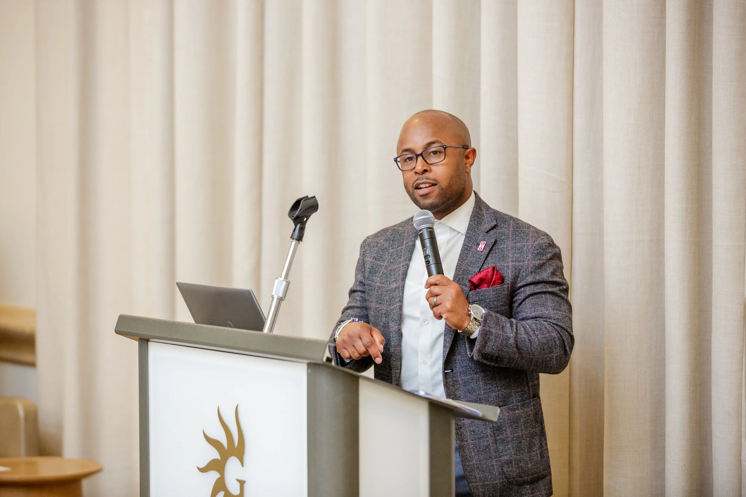 This is Dr. Dexter Shorter in a gray plaid blazer, white shirt, and glasses stands at a podium, speaking into a microphone during a presentation or speech.