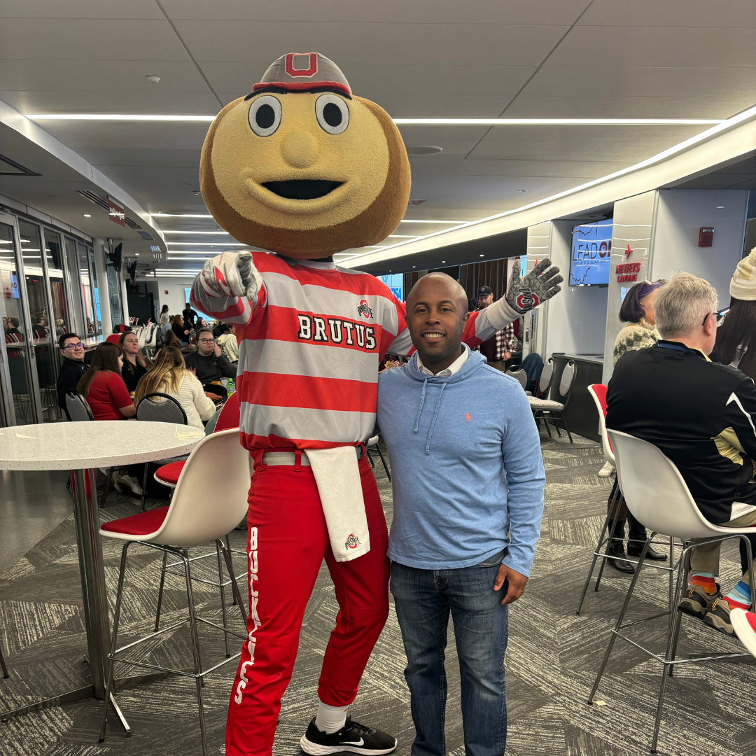 This is Dr. Dexter Shorter standing next to a person in the Ohio State University mascot costume that says 'BRUTUS' written on it, in an indoor setting with seated people in the background.