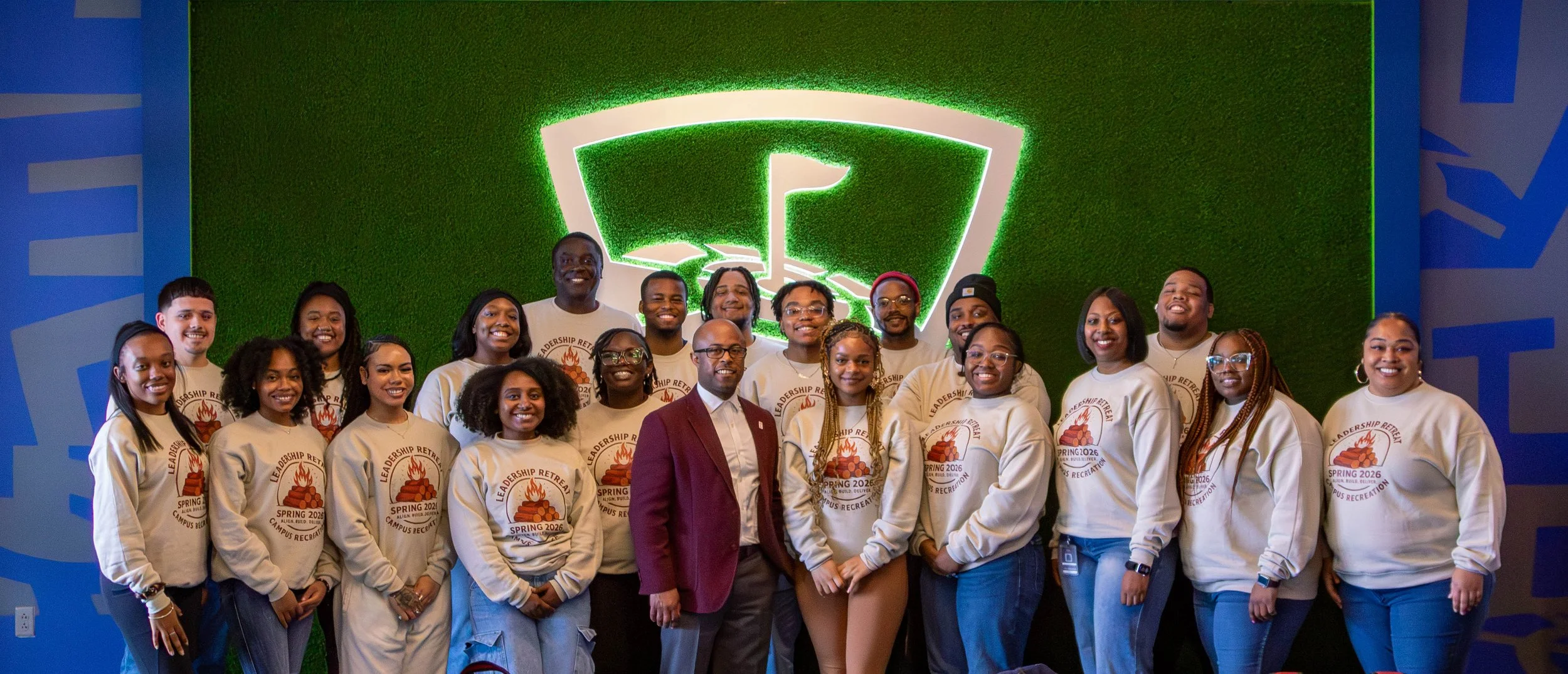 This is Dr. Dexter Shorter with young adults posing for a photo in front of a green wall with a logo featuring a flame, some wearing white sweatshirts with a logo and text 'Leadership Retreat Spring 2026 Campus Recreation'.