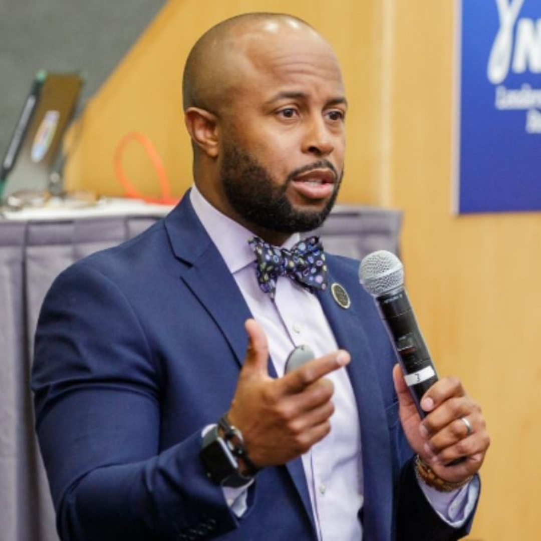 This is Dr. Dexter Shorter in a blue suit with a bow tie speaking into a microphone at a conference or event.