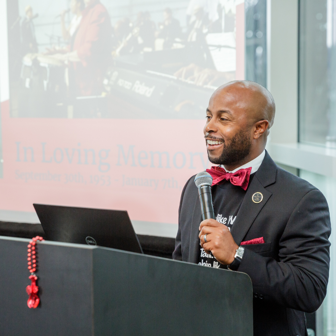 This is Dr. Dexter Shorter in a suit with a red bow tie speaking into a microphone an event, with a presentation slide in the background.