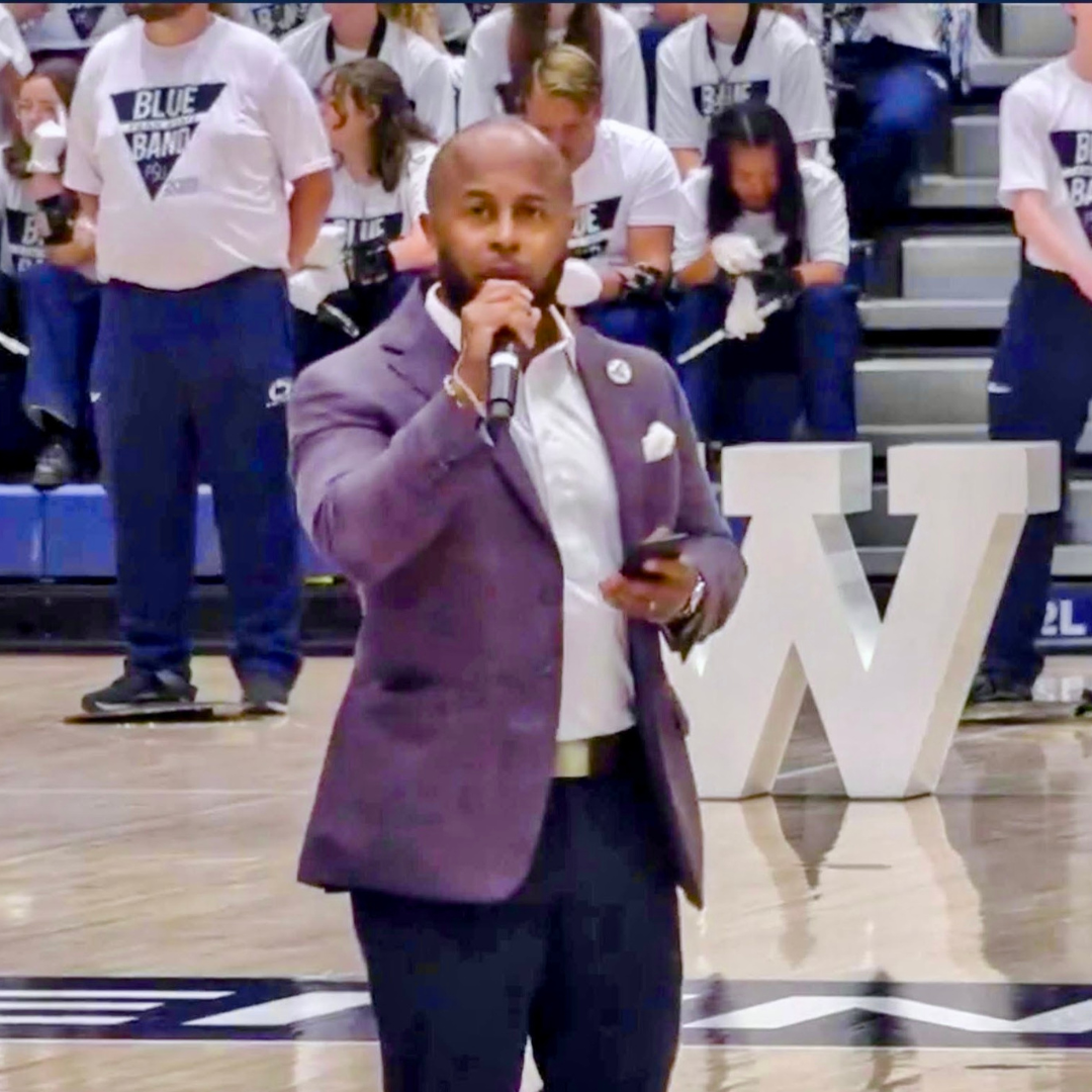 This is Dr. Dexter Shorter in a purple suit jacket, white shirt, and dark pants speaking into a microphone on a basketball court. Behind him, a group of Penn State students in white t-shirts with a blue logo sits on bleachers.