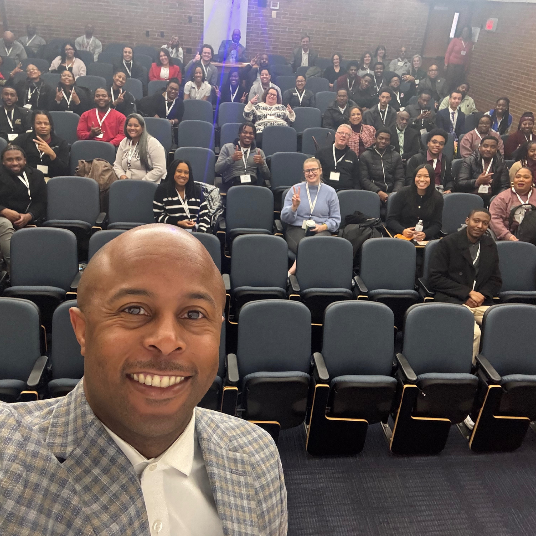 This is Dr. Dexter Shorter in a checkered blazer taking a selfie in front of a large group of diverse people seated in a conference room with blue chairs, brick walls, and a brick wall background.