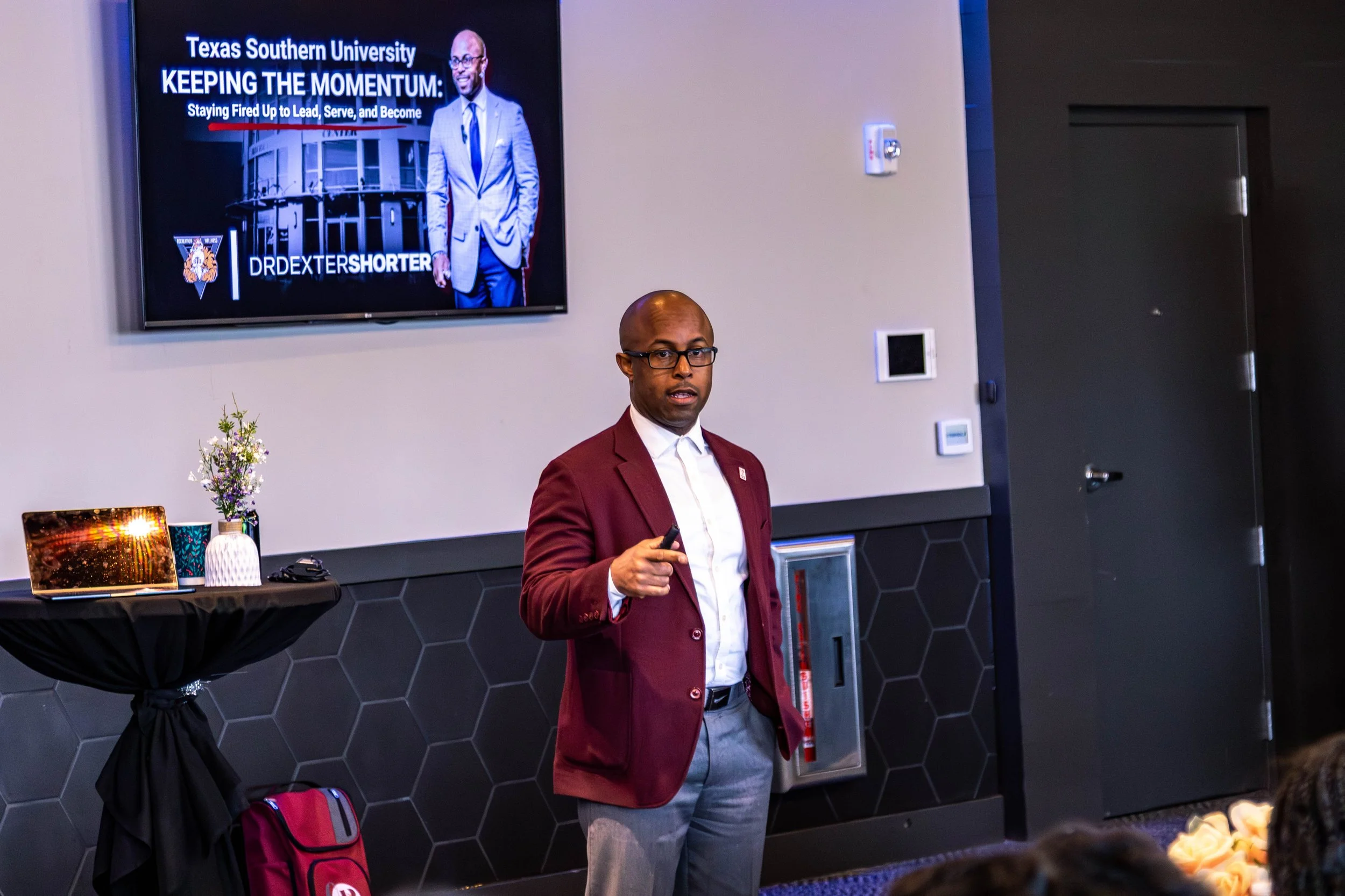 This is Dr. Dexter Shorter in a maroon blazer and glasses giving a presentation to a group of students in a conference room. A large screen behind him displays a slide with the title 'Texas Southern University' and 'Keeping the Momentum'.