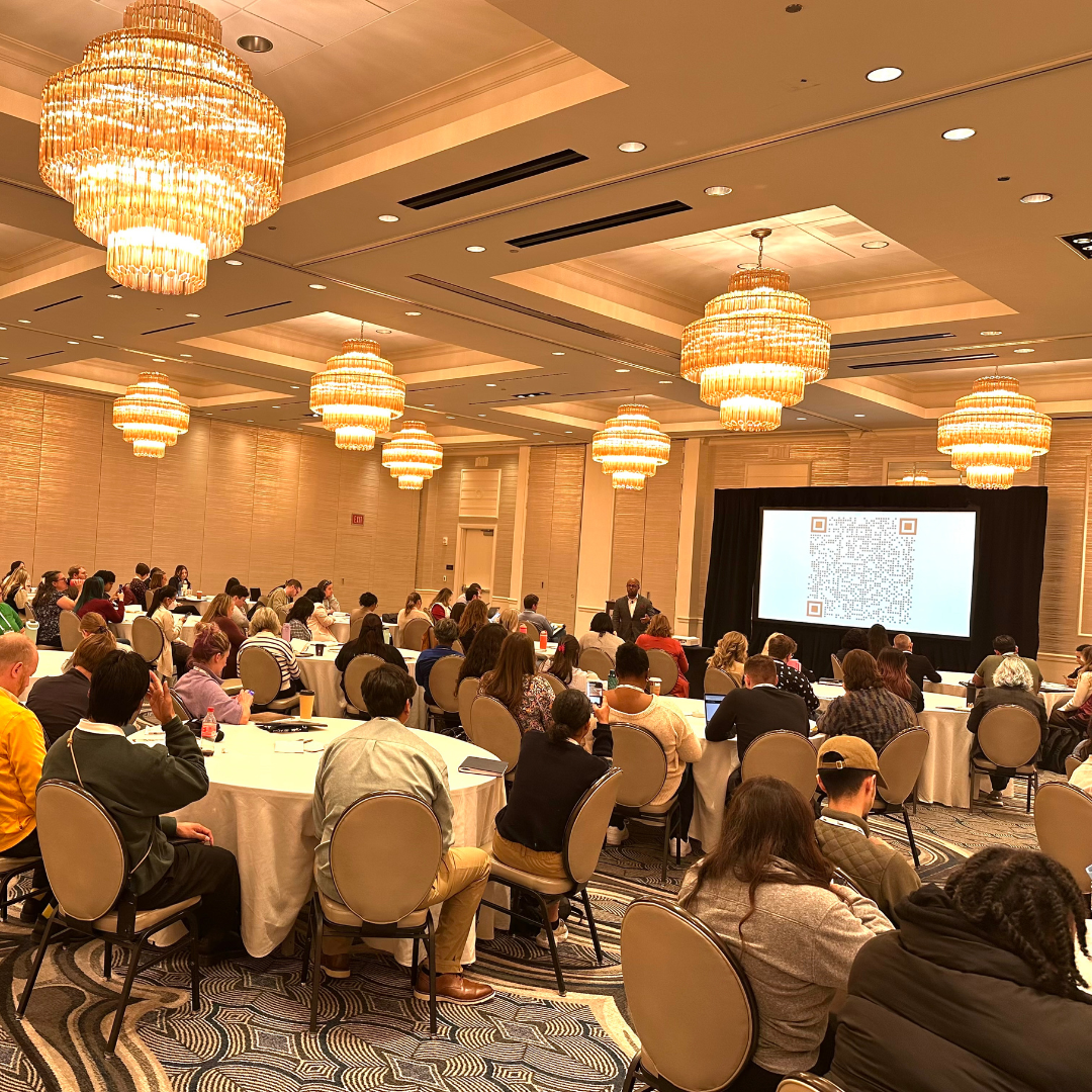 This is Dr. Dexter Shorter delivering a keynote speech to attendees sitting at round tables in front of a large screen. Multiple chandeliers hang from the ceiling, illuminating the room.