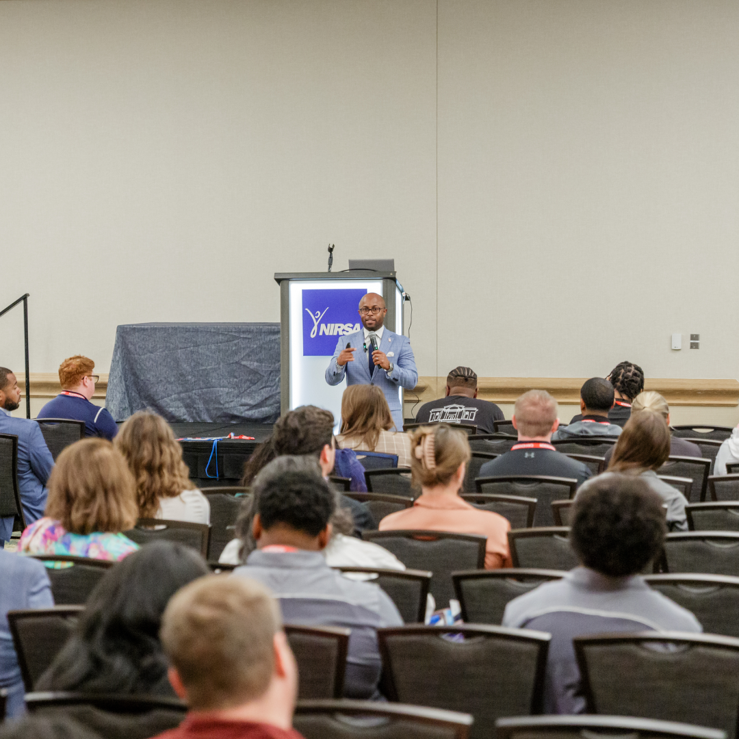 This is Dr. Dexter Shorter in a blue checkered  blazer speaking at a conference while holding a microphone with a presentation slide behind him displaying the NIRSA logo. Audience seated in a conference room listening.