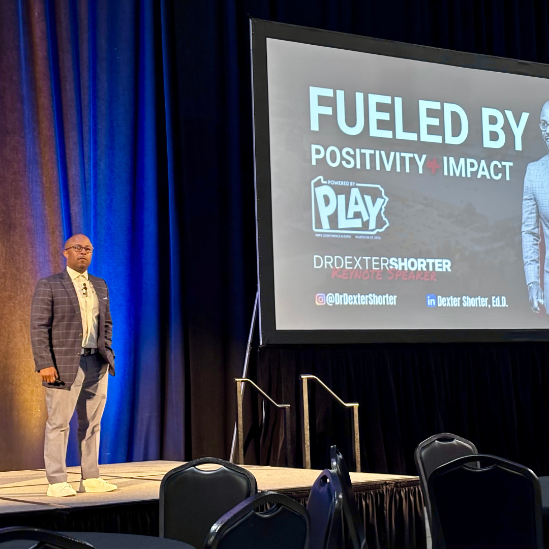 This is Dr. Dexter Shorter standing on a stage giving a presentation at an expo, with a large screen displaying the title "FUELED BY POSITIVITY + IMPACT" and his social media handles.