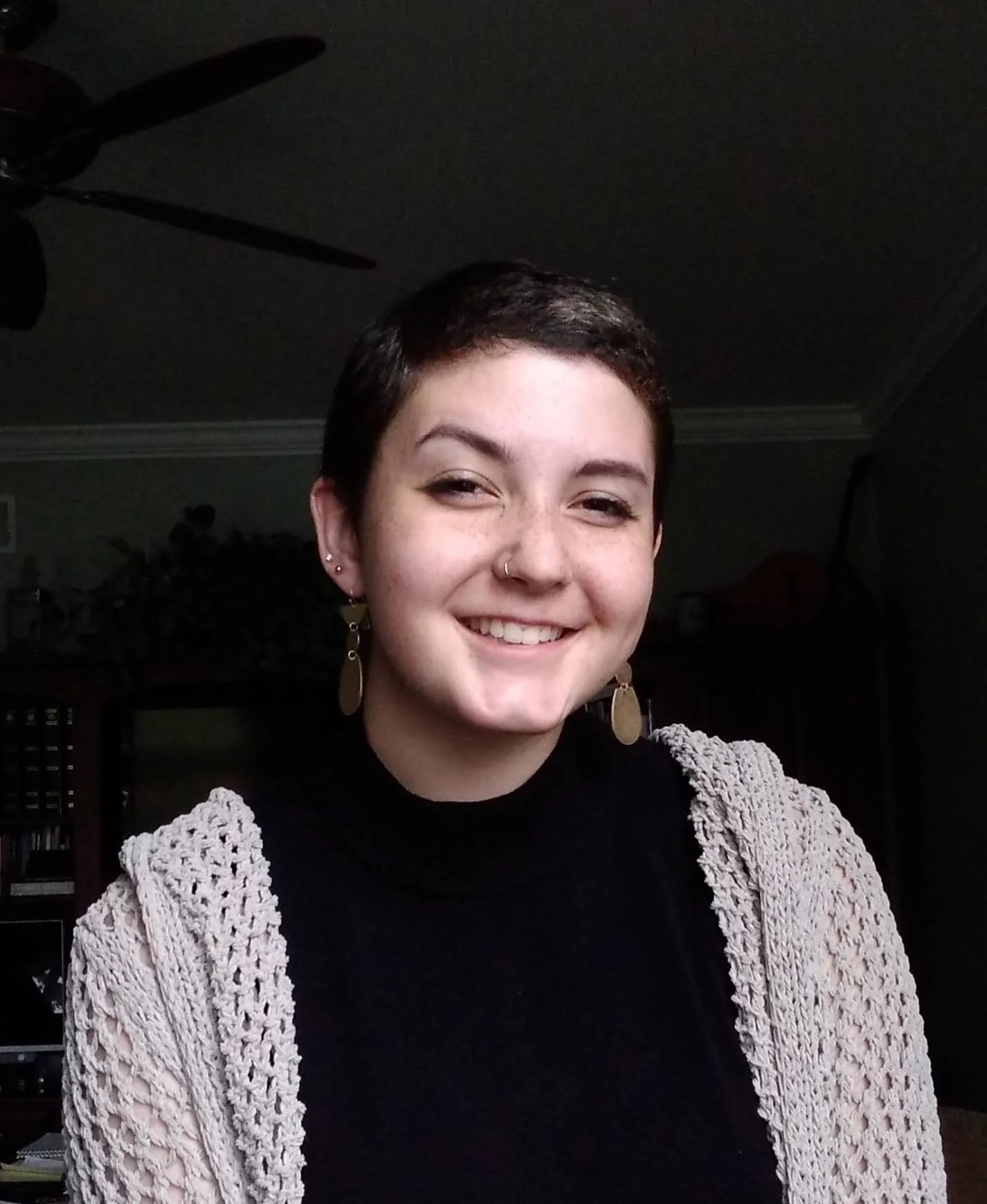 A young woman with short dark hair, wearing a black top and beige crochet cardigan, smiling in a home setting with dark ceiling and a ceiling fan in the background.