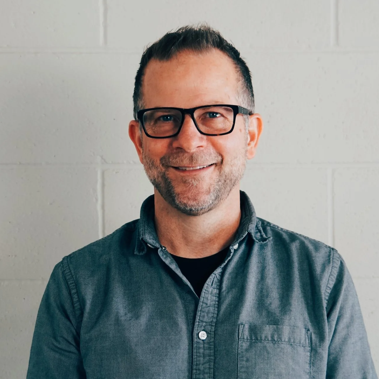 Smiling man with glasses and a beard wearing a gray button-up shirt, standing against a white brick wall.