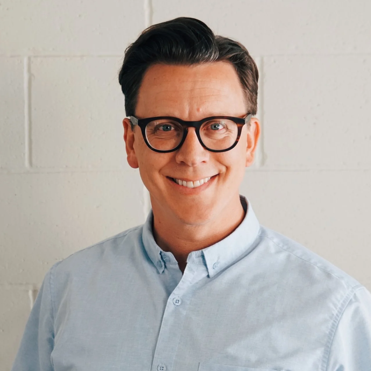 Person wearing glasses and a light blue shirt smiling against a white brick wall.