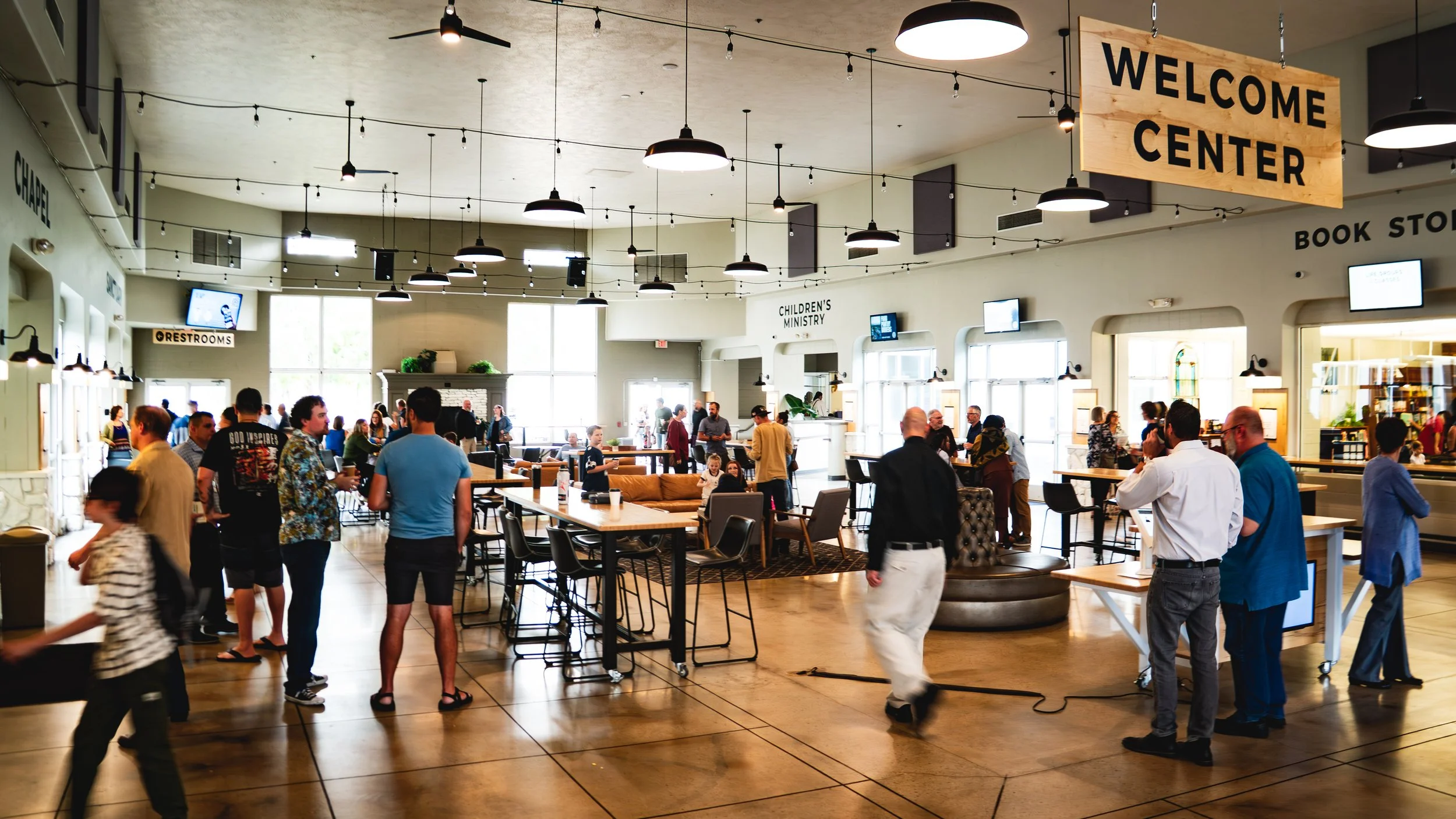 A group of people having a conversation indoors, with a coffee bar visible in the background. The setting appears casual and cozy, with framed decorations on the walls and a counter with coffee supplies.