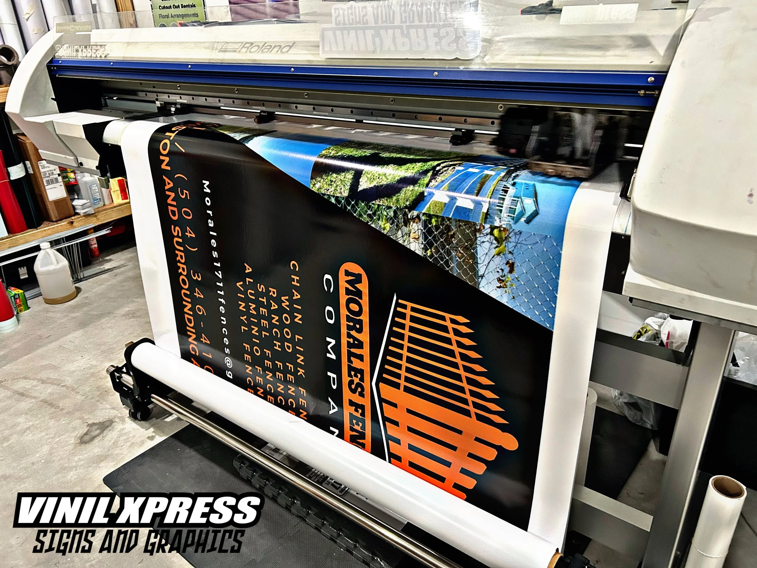 A large poster printing machine with a printed banner featuring an orange and black logo for Morale Storm, along with a list of locations and contact info, in a workshop setting.