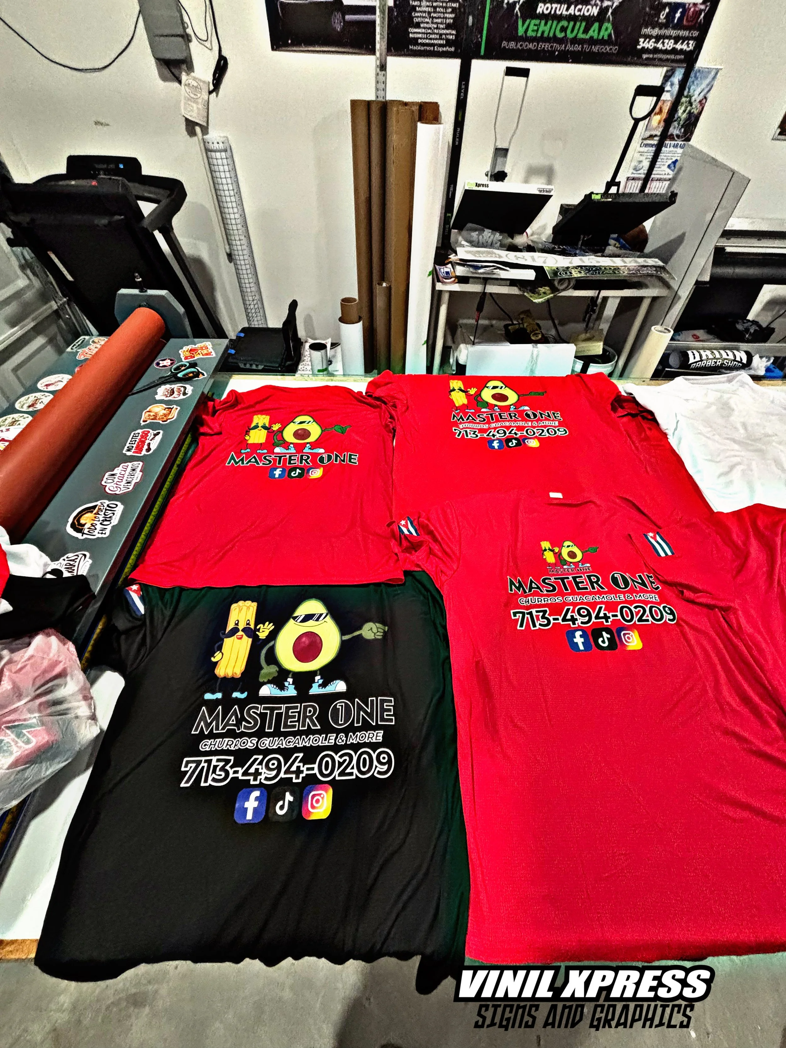Red and black T-shirts for 'Master One' restaurant, featuring cartoon avocado and churros characters, contact number, and social media icons, lying on a workshop table with vinyl cutting tools and rolled materials in the background.
