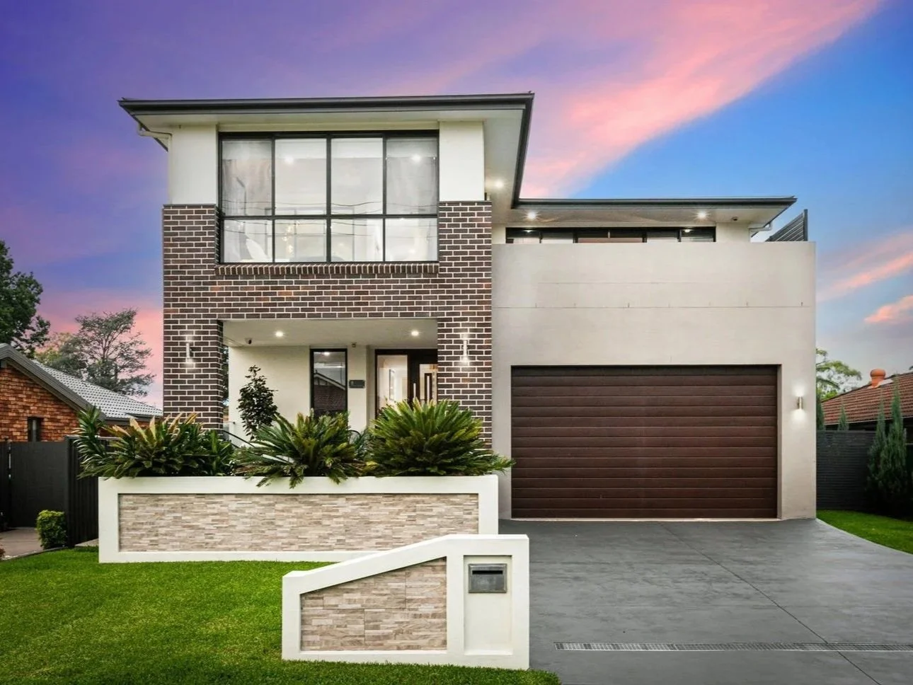 Modern two-story house with brick and concrete exterior, large front window, landscaped front yard, and attached garage, during sunset.