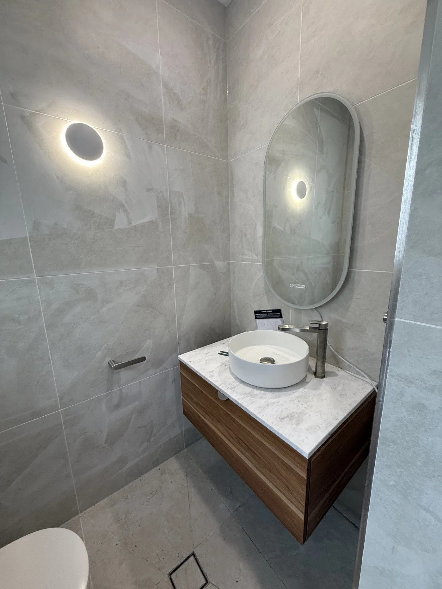 Modern bathroom sink with a round vessel basin, a tall faucet, oval mirror, grey tiled walls, and a wall-mounted grab bar.