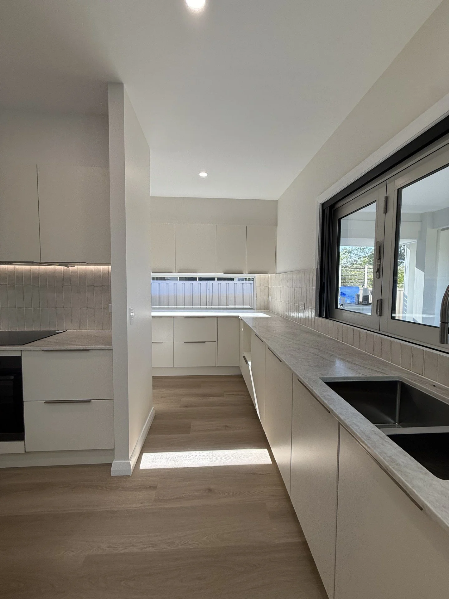 Modern kitchen with white cabinets, countertops, and a black sink, illuminated by natural light through large windows.