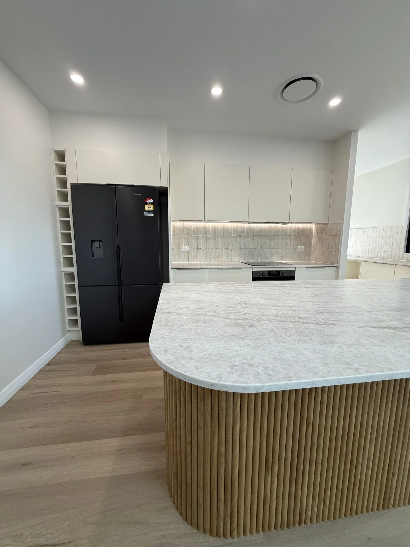 Modern kitchen with white cabinets, black refrigerator, beige tiled backsplash, and a large curved marble-topped island with a wooden slat base.