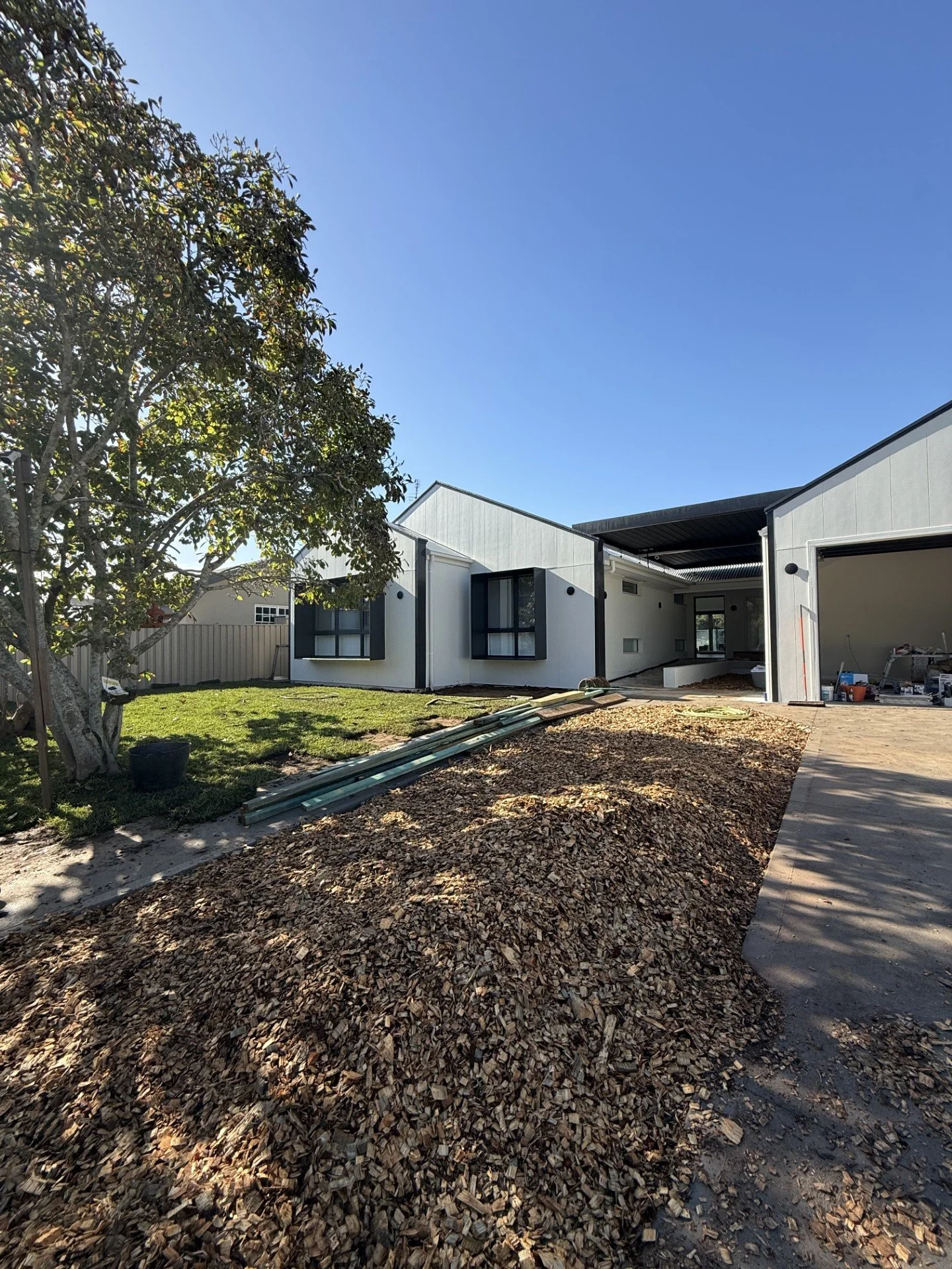 Modern house under construction with new landscaping, a tree on the left, and a clear blue sky.
