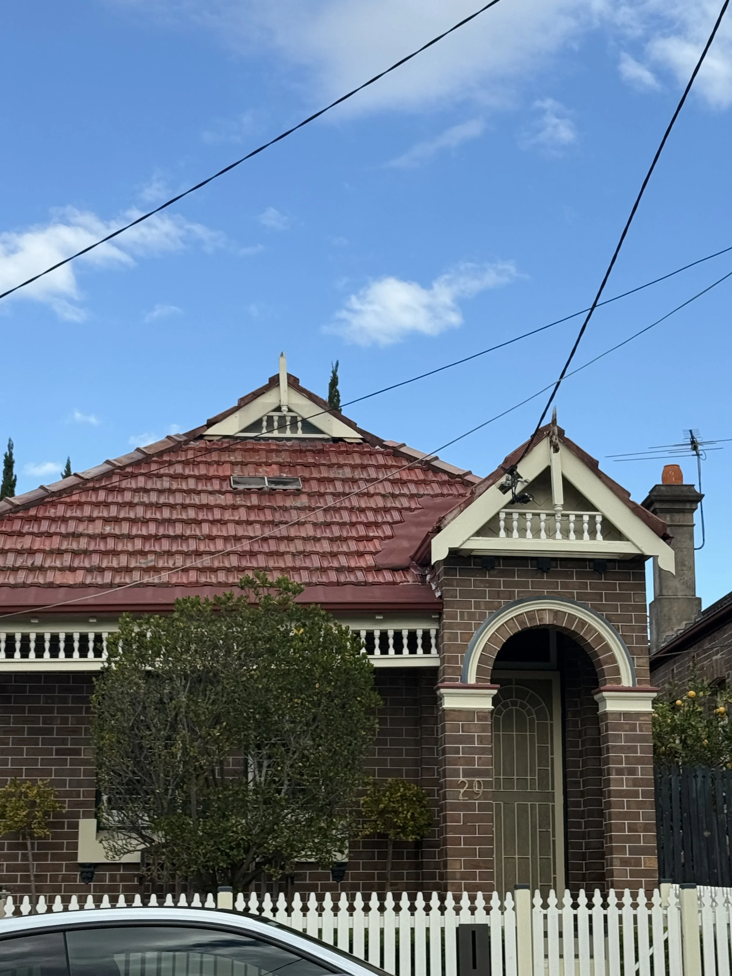 A brick house with a red tiled roof, a white picket fence, and a tree in front. The house has decorative white trim and an arched entryway.