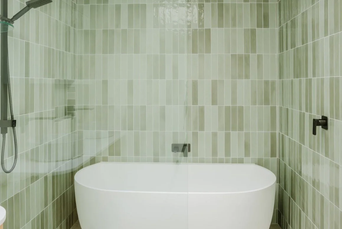 Modern bathroom with a white freestanding bathtub against a green tiled wall, a showerhead on the left, and a glass partition.