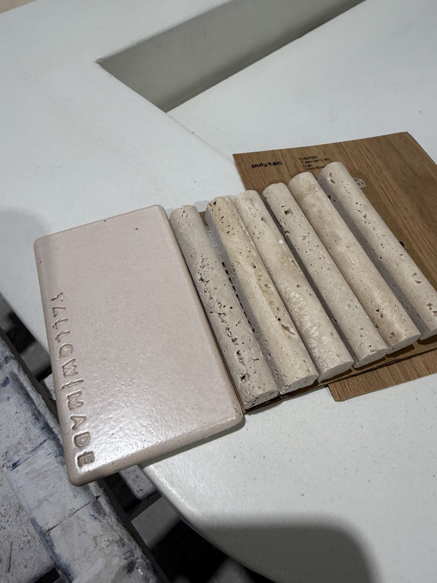 A sample display of tile materials, including a rectangular tile with the word 'MOPIN' embossed on it, several cylindrical stone tiles in varying shades and textures, and a wooden sample under them, all arranged on a white surface.