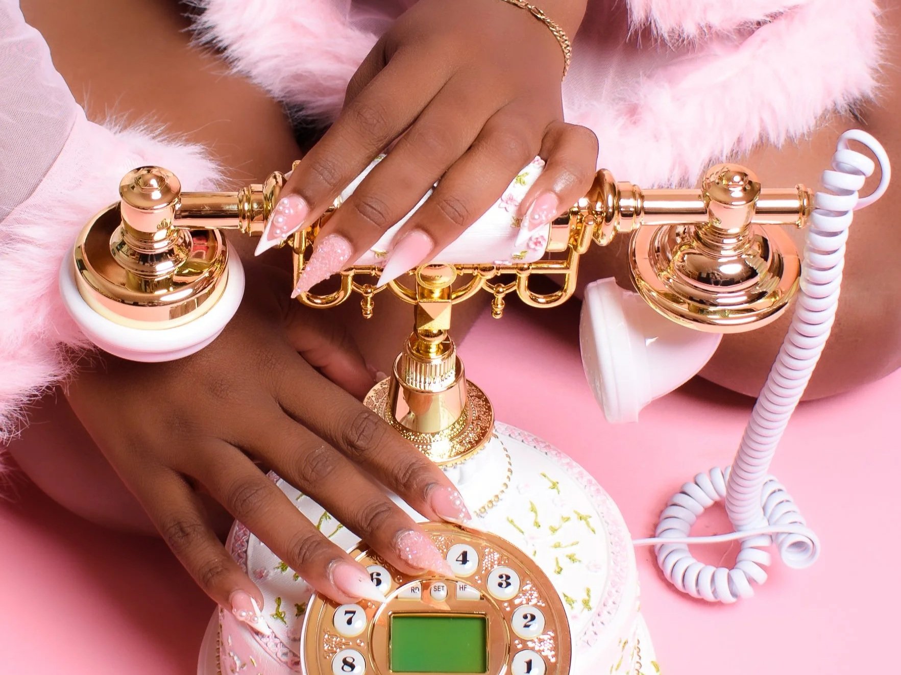 Close-up of a person with decorated nails pressing buttons on a vintage rotary phone with a pastel pink and gold design, resting on a matching pink surface with fuzzy pink fabric in the background.