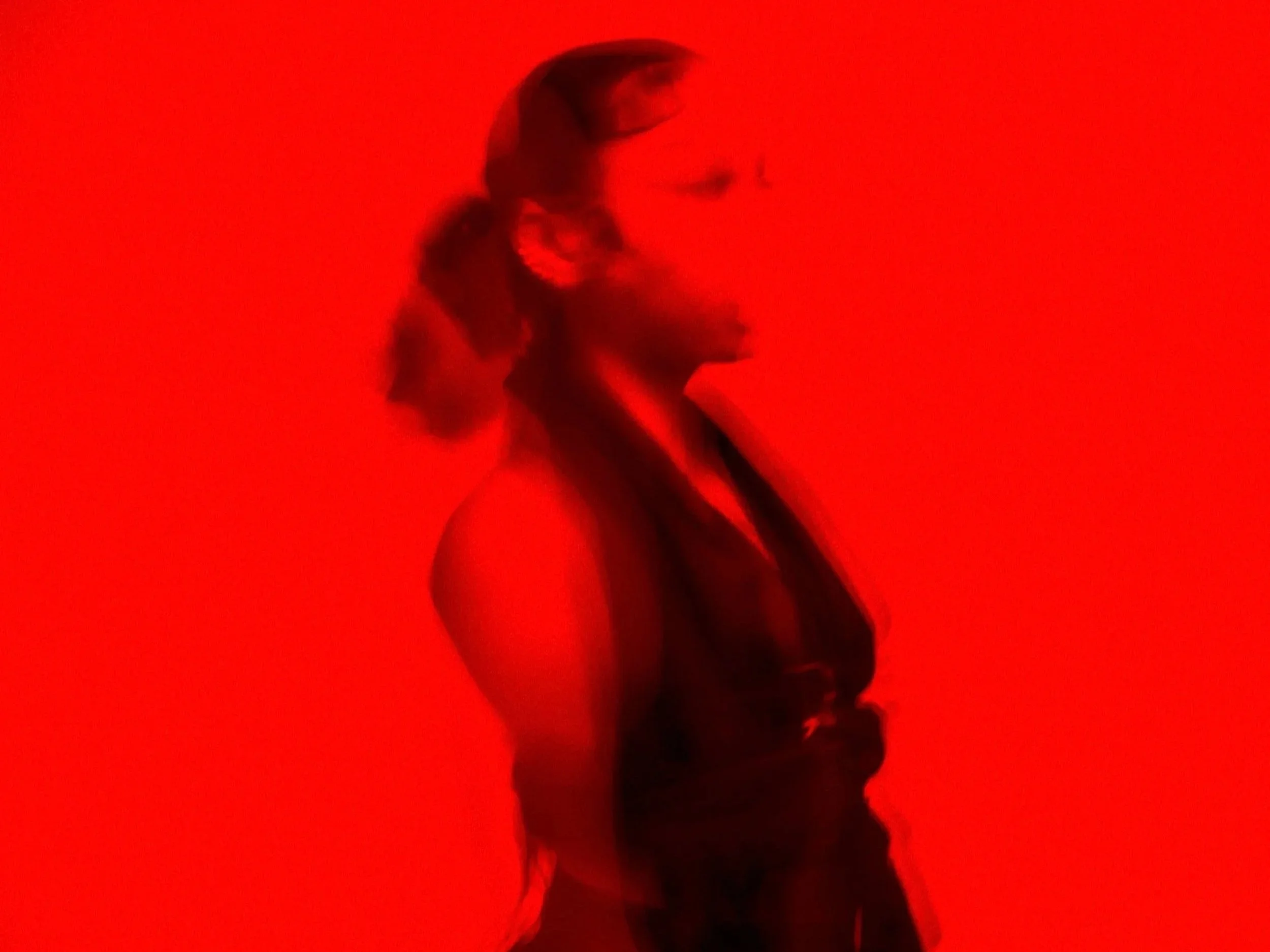 A woman with long hair tied back, wearing a sleeveless top, stands in front of a bright red background.