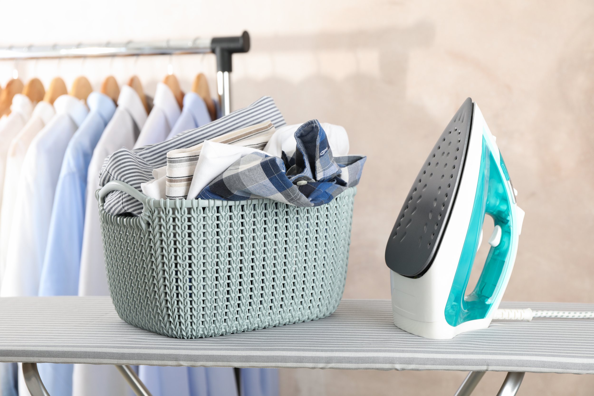 A laundry basket filled with folded and unfolded clothes next to a steam iron on a striped ironing board.