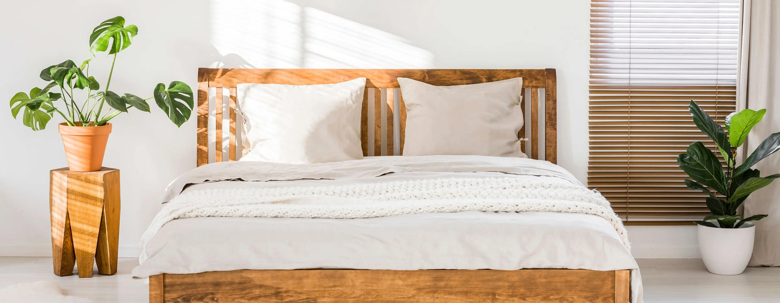 A bedroom with a wooden bed frame, cream-colored bedding, and two earth-toned pillows. There are two potted plants, one on a wooden stand on the left and another in a white pot on the right, with sunlight streaming through a window with blinds.