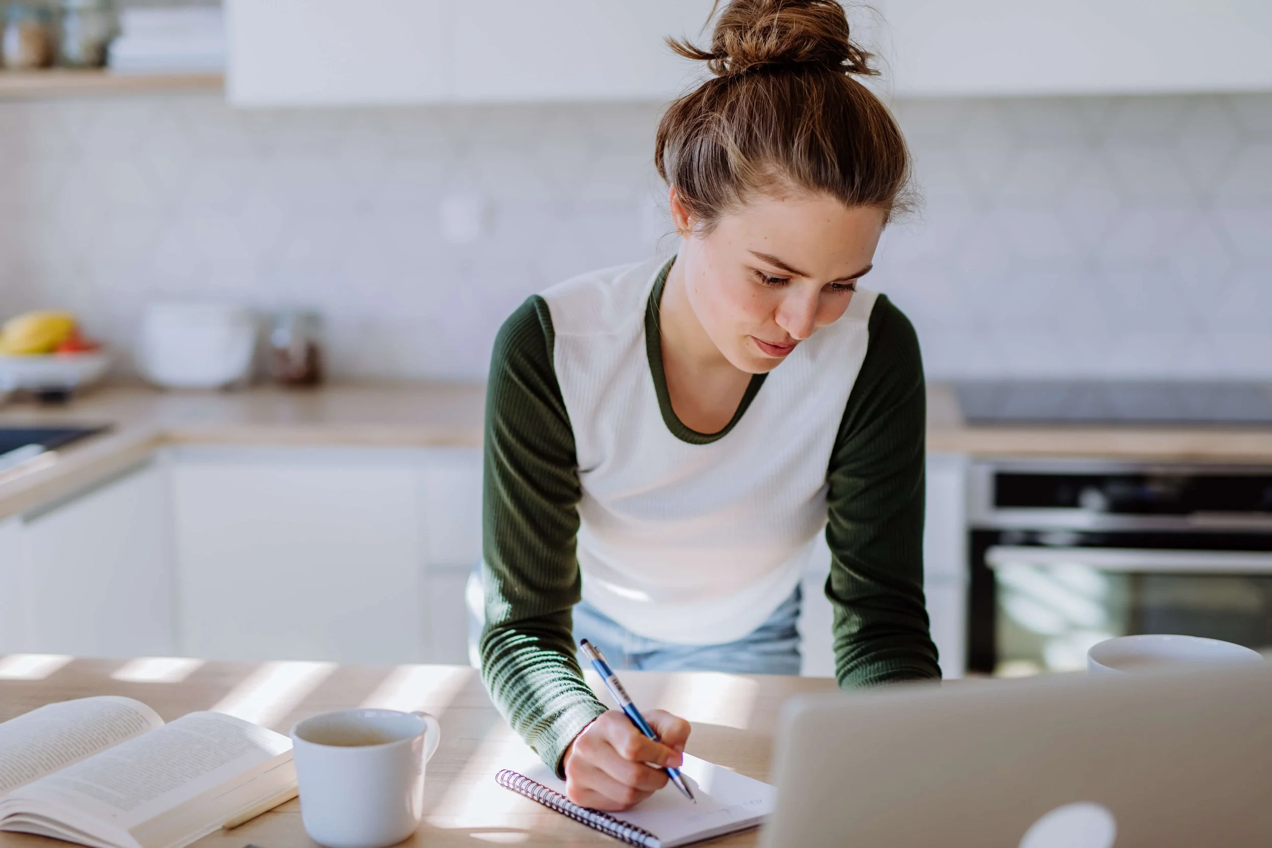A young woman with brown hair tied in a bun, wearing a white and green long sleeve shirt, is writing in a notebook on a kitchen counter. There is a cup of coffee and an open book on the counter, with a laptop in front of her. The background shows a modern kitchen with white cabinets and a gray backsplash.
