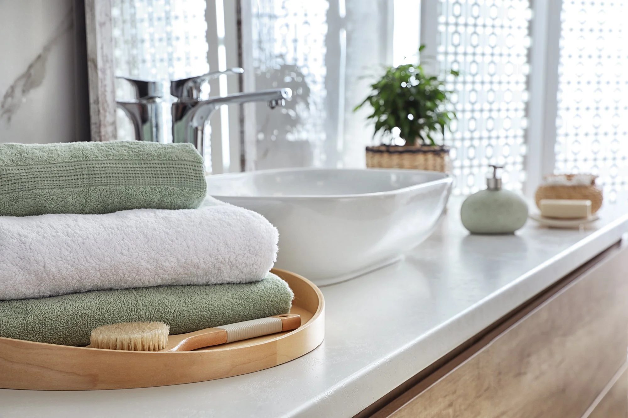 Stacked green and white towels on a round wooden tray with a brush, on a bathroom countertop near a sink with a silver faucet.