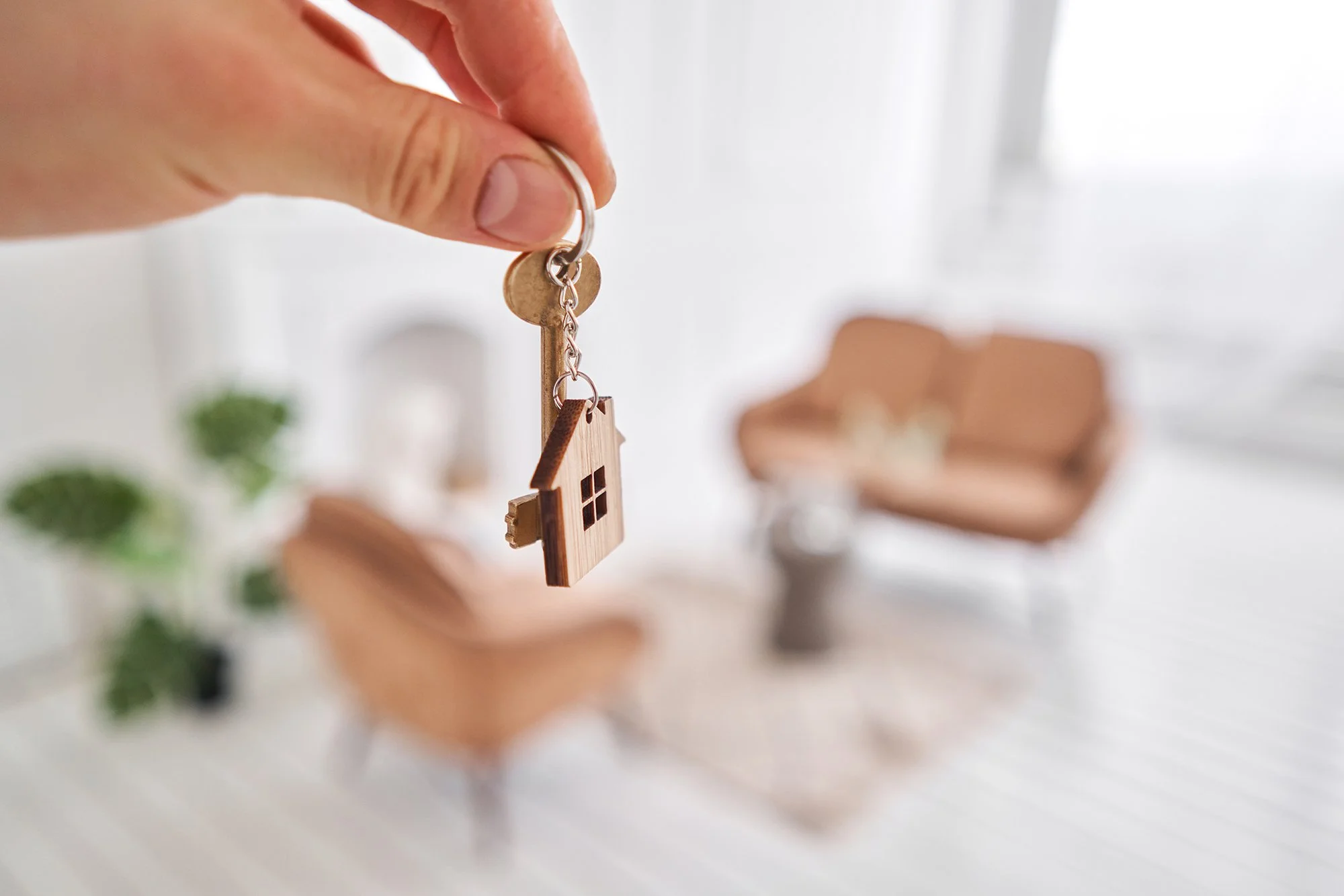 Close-up of a hand holding a keychain with a small wooden house charm in the foreground. The background shows a blurred living room with a chair and some greenery.