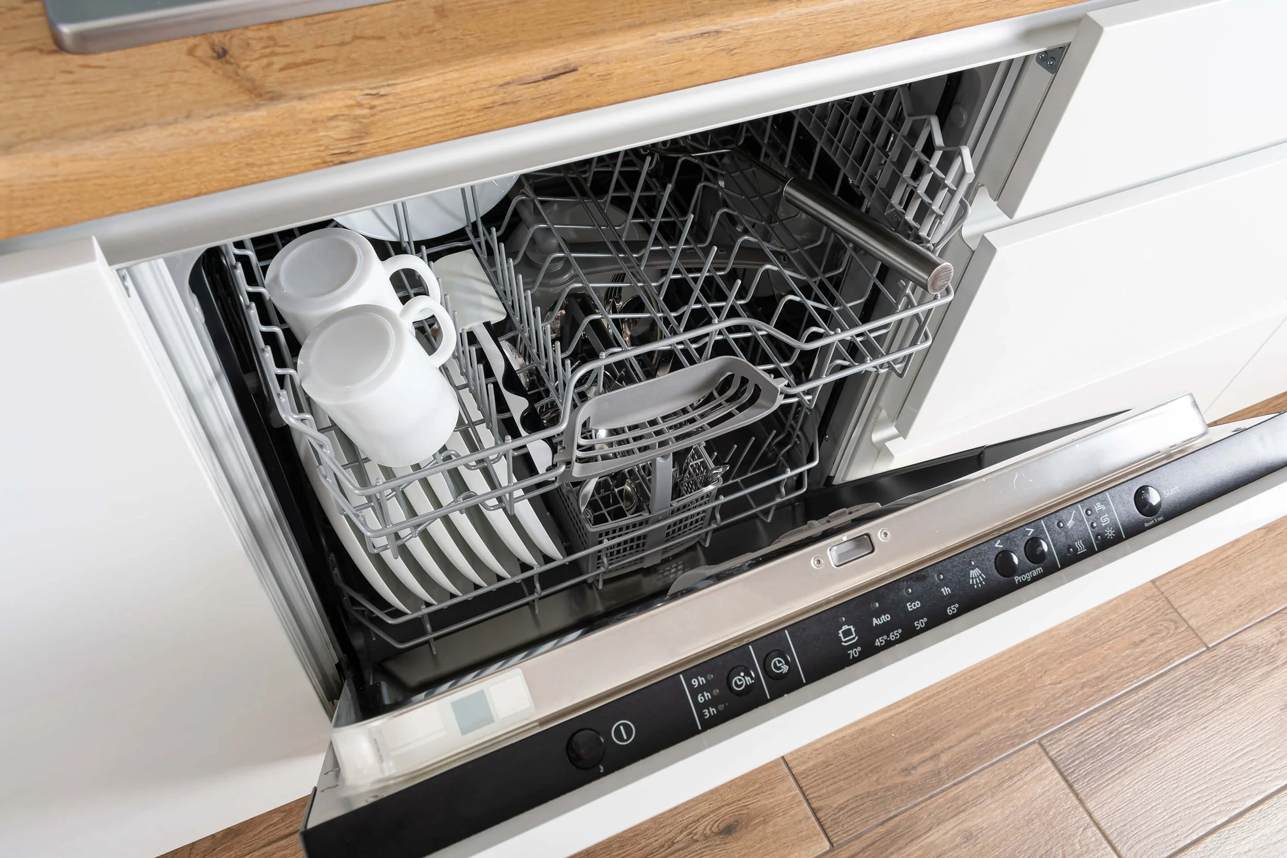 Open dishwasher filled with white ceramic mugs and utensils, with a wooden countertop above and a control panel visible at the bottom.