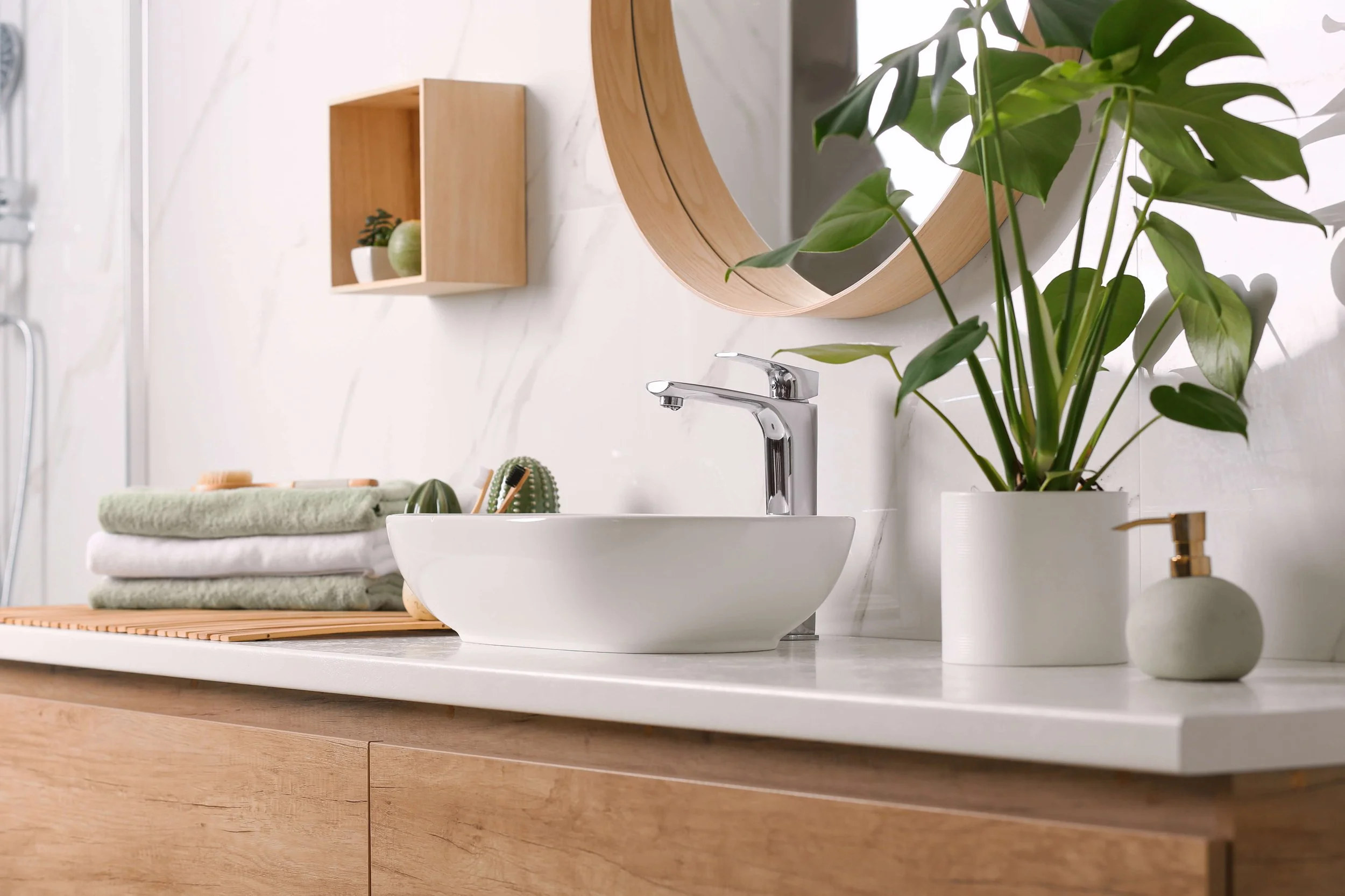 A modern bathroom sink area with a white vessel sink, a silver faucet, and green plants in white pots. There are folded towels on a wooden tray and a round wooden mirror on the wall.