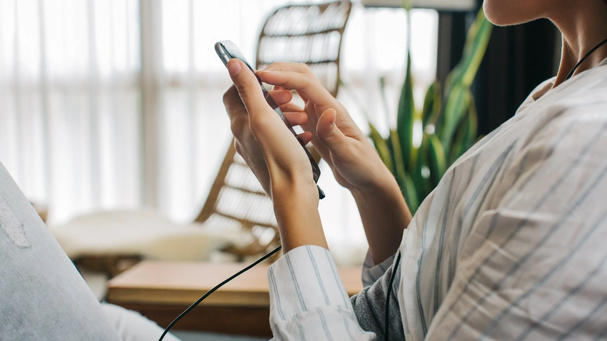 A person using a smartphone with earphones in a bright indoor setting, possibly a home or cafe, with a wooden chair and green plant in the background.