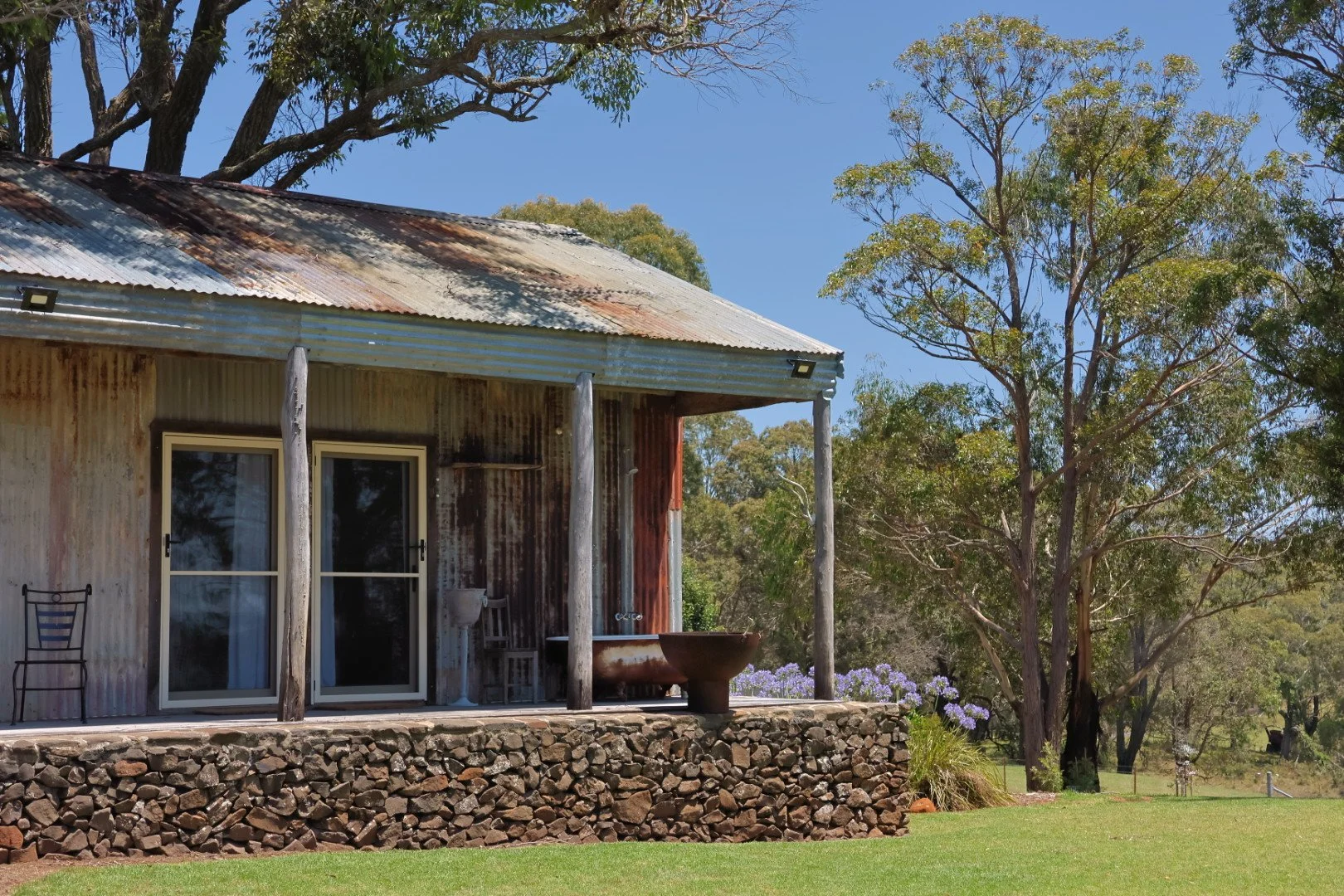 A rustic corrugated metal house with a porch, surrounded by green grass, trees, and purple flowers, under a clear blue sky.