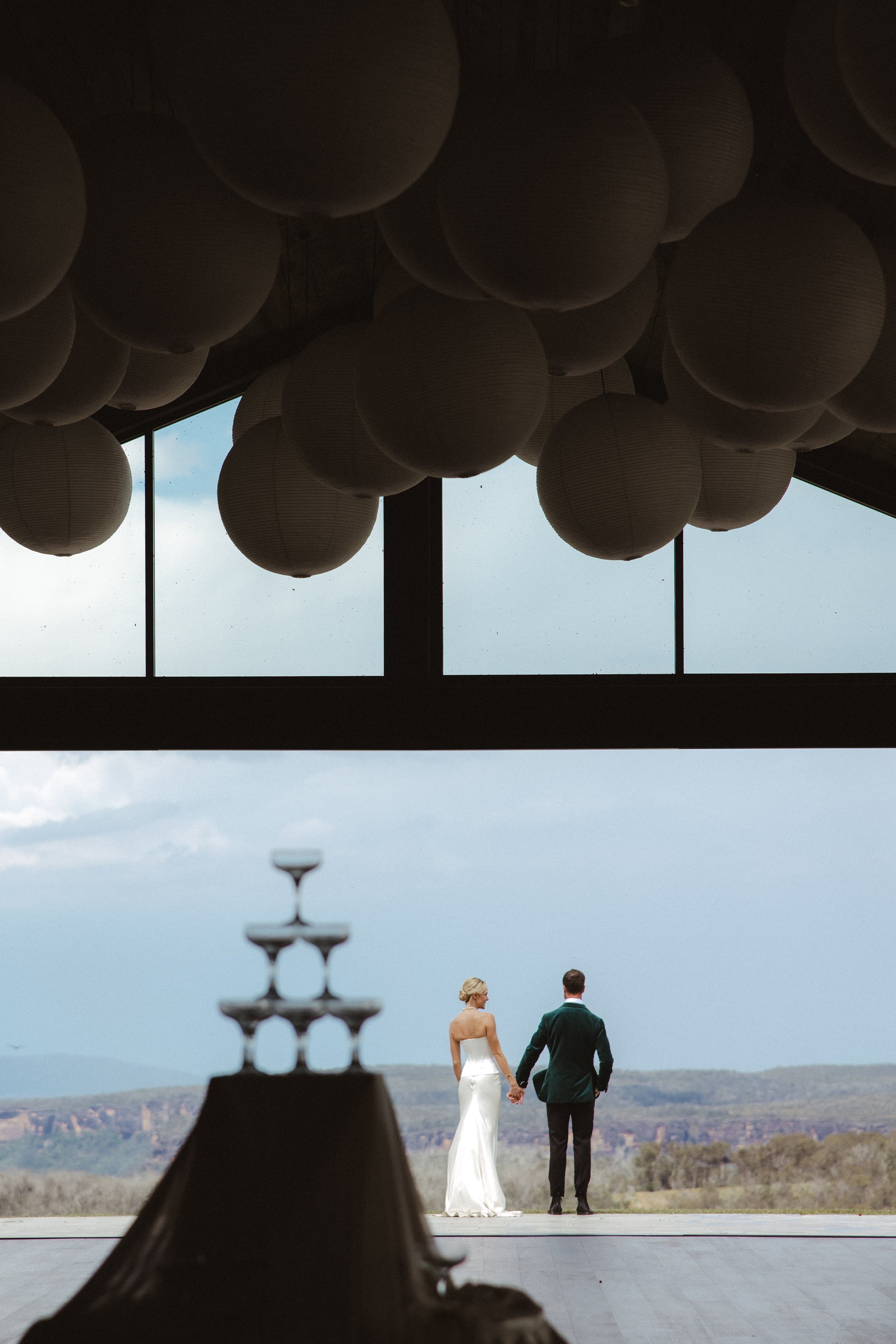 A bride and groom holding hands, standing outdoors with a scenic landscape in the background, viewed from inside a building with hanging paper lanterns and a decorative fountain in the foreground.
