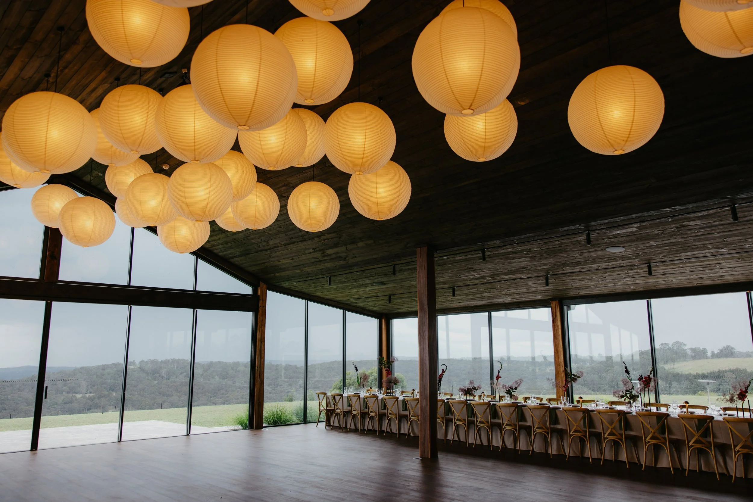 Interior of a modern event space with hanging paper lanterns, large glass windows revealing a scenic outdoor landscape, and a long table set for a meal with floral centerpieces.