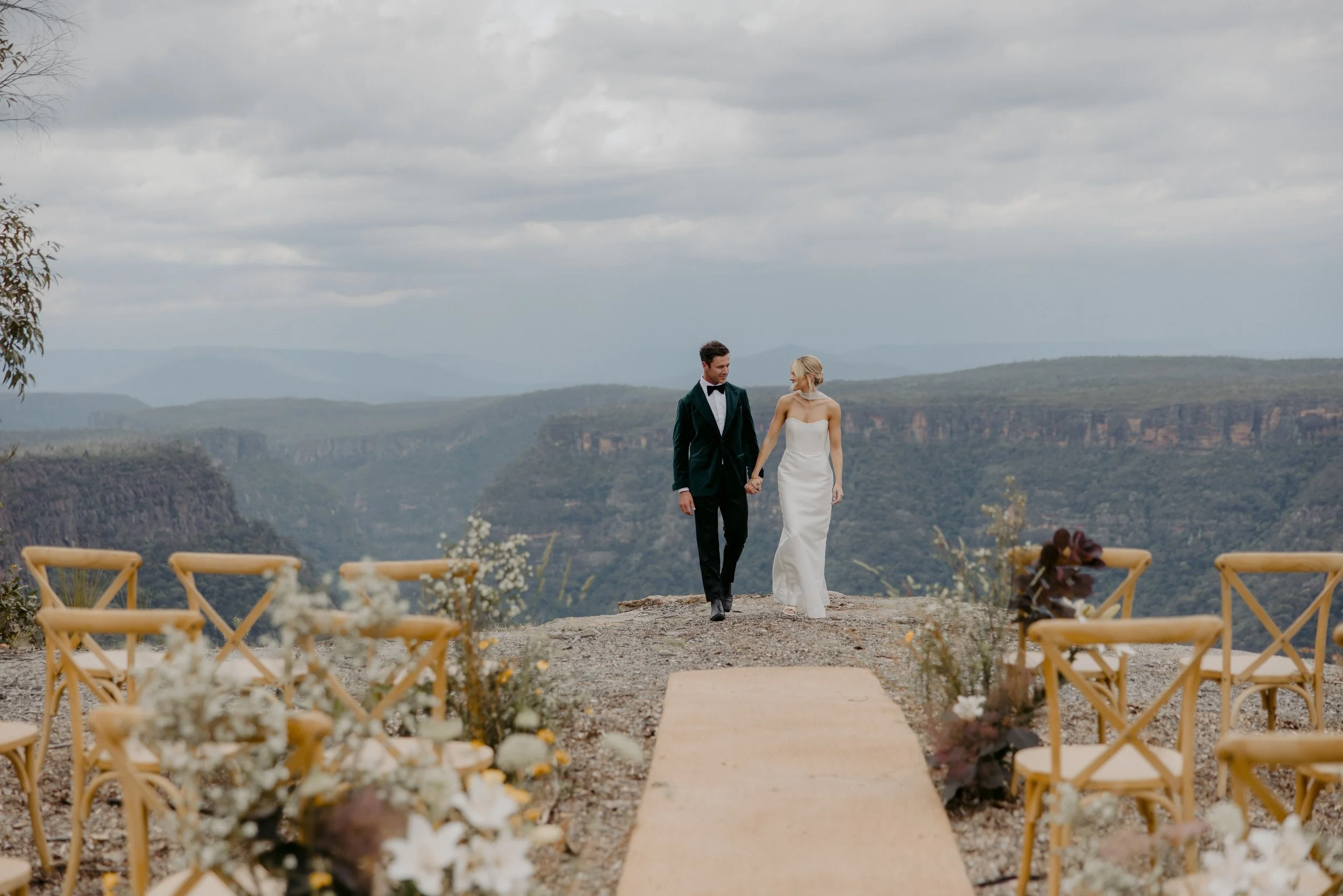 A bride and groom holding hands while walking down an outdoor aisle with scenic canyon views in the background, decorated with flowers and chairs.