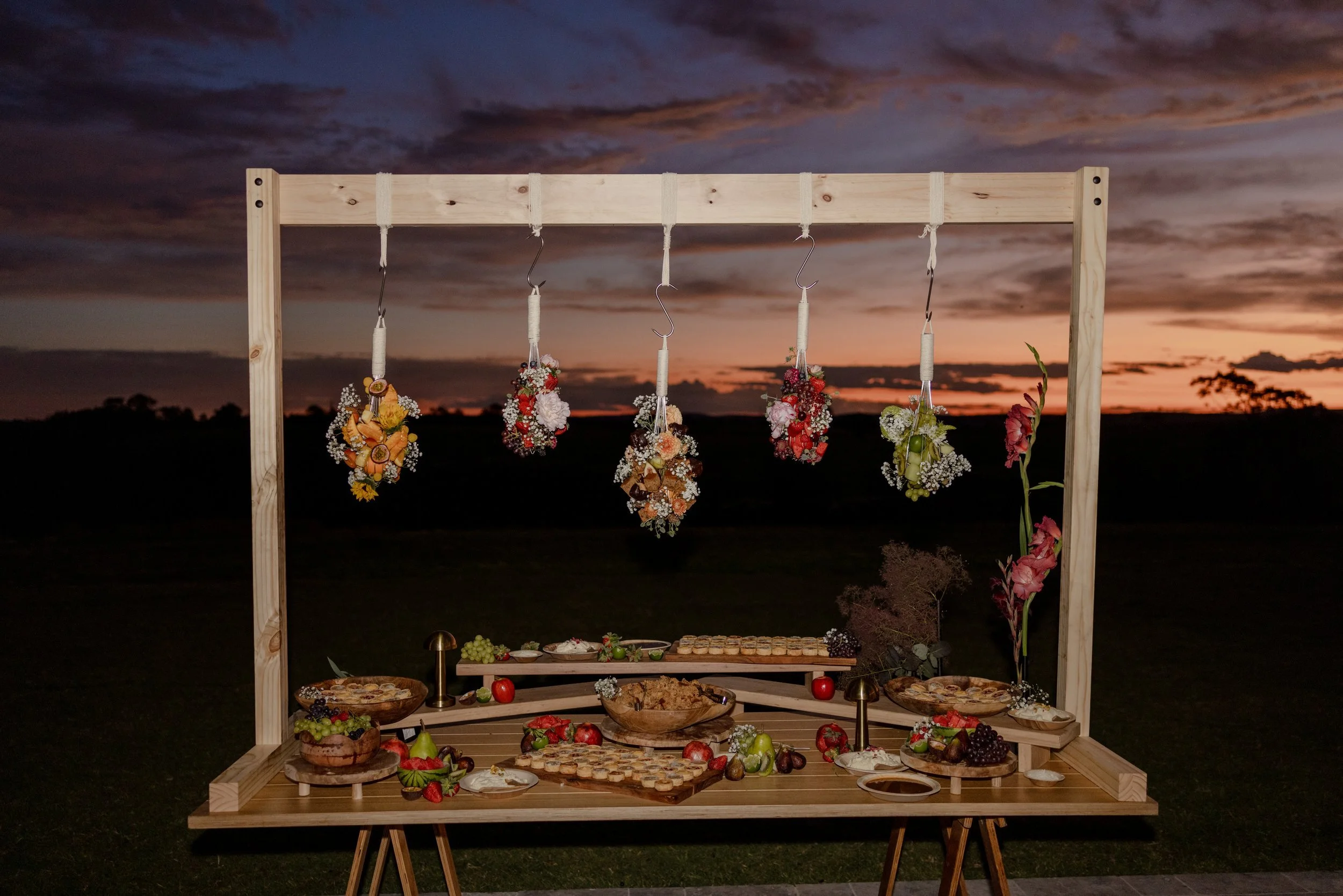 A nighttime outdoor scene with a decorated wooden frame displaying hanging floral arrangements, and a table with an assortment of food, fruits, and flowers, set against a sunset sky with dark clouds.