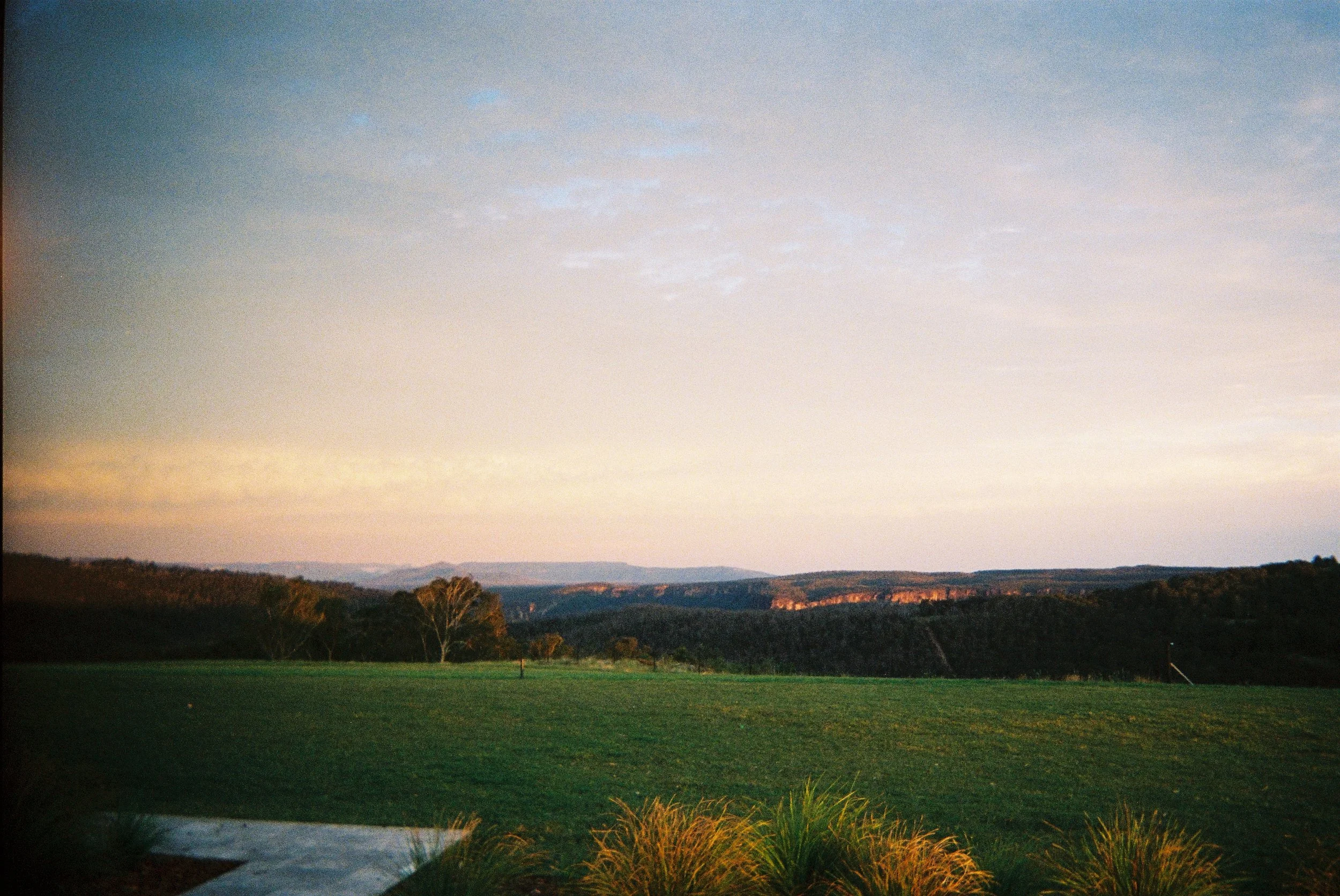 A landscape scene at sunset with a grassy field in the foreground, a few trees, distant hills, and a partly cloudy sky.