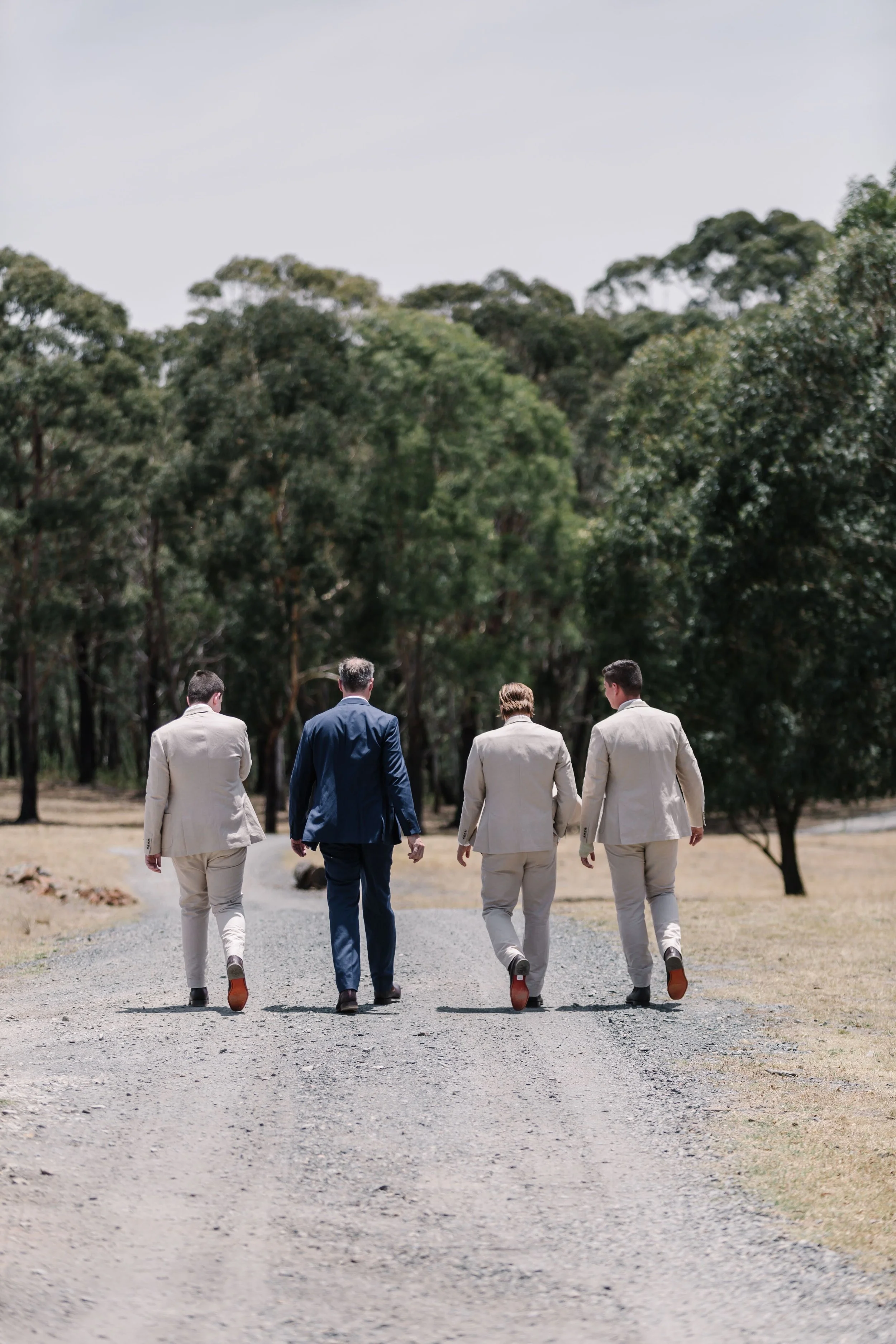 Four men walking on a gravel path through a forest, dressed in suits, with three in beige suits and one in a blue suit.