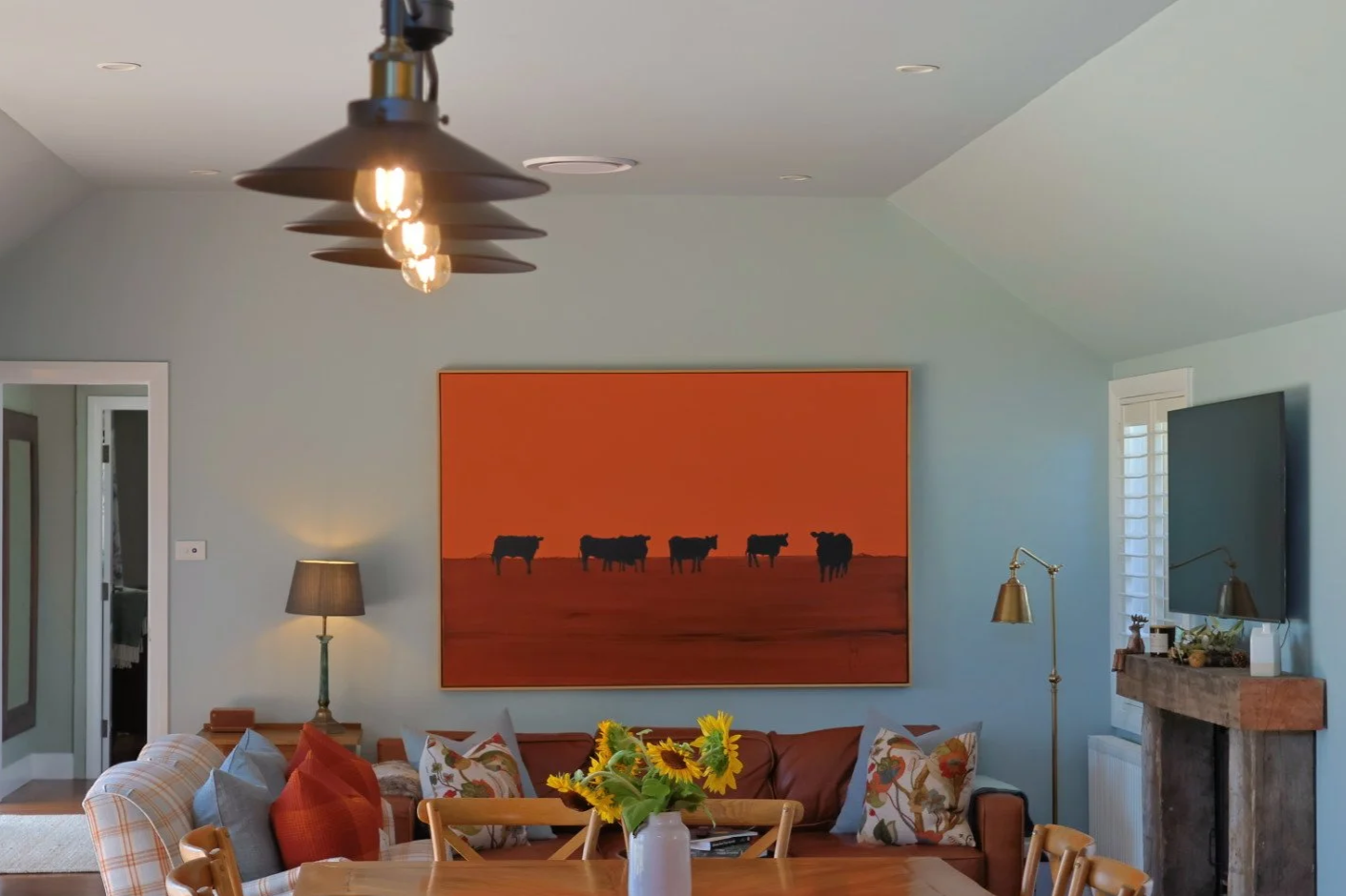 Living room with a large orange painting of cows, a brown leather sofa with pillows, a wooden dining table with a sunflower vase, a fireplace, and a wall-mounted TV.