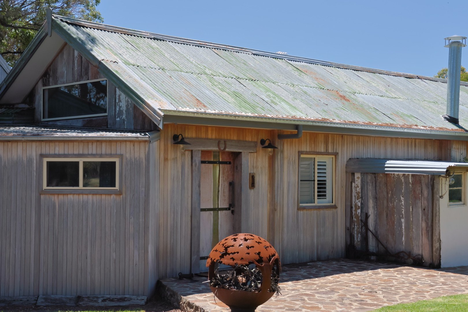 Old rustic house with weathered metal roof, two small windows, and a decorative firepit with black silhouettes of bats, situated on a stone patio.