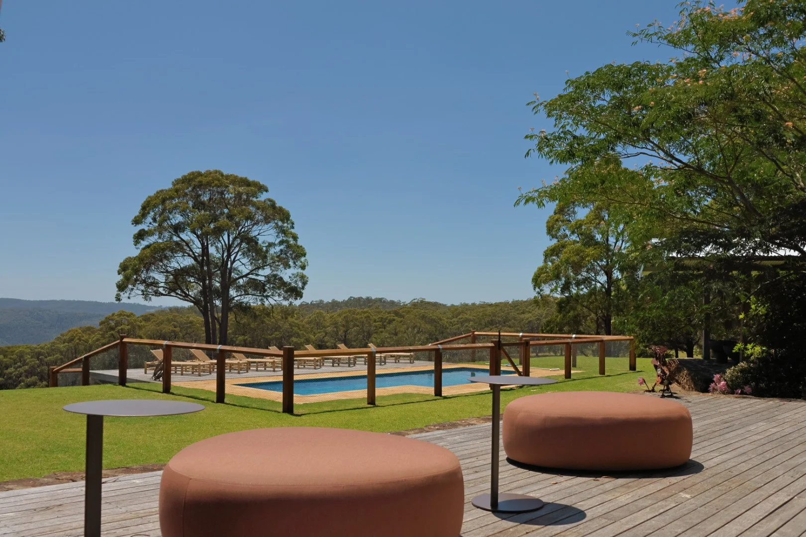 A patio with two large round cushioned seats and small round tables, overlooking a fenced swimming pool surrounded by green grass and large trees with a mountain view in the background.