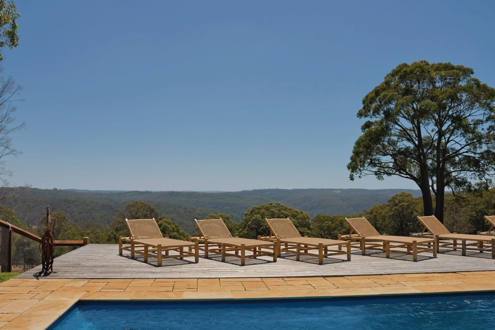 Outdoor view of lounge chairs by a pool on a wooden deck overlooking a landscape with trees and distant hills under a clear blue sky.