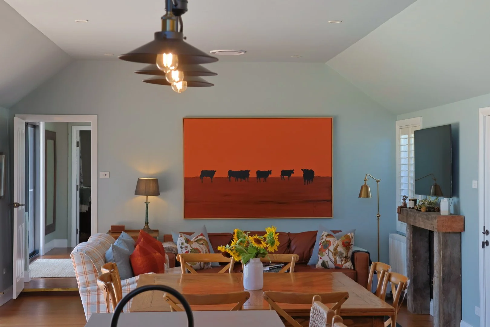 Living room with a wooden dining table and chairs, a colorful sofa with throw pillows, a large orange painting with cows, a TV on a mounted wall, and a fireplace with decor. There is a large window and a door leading outside.