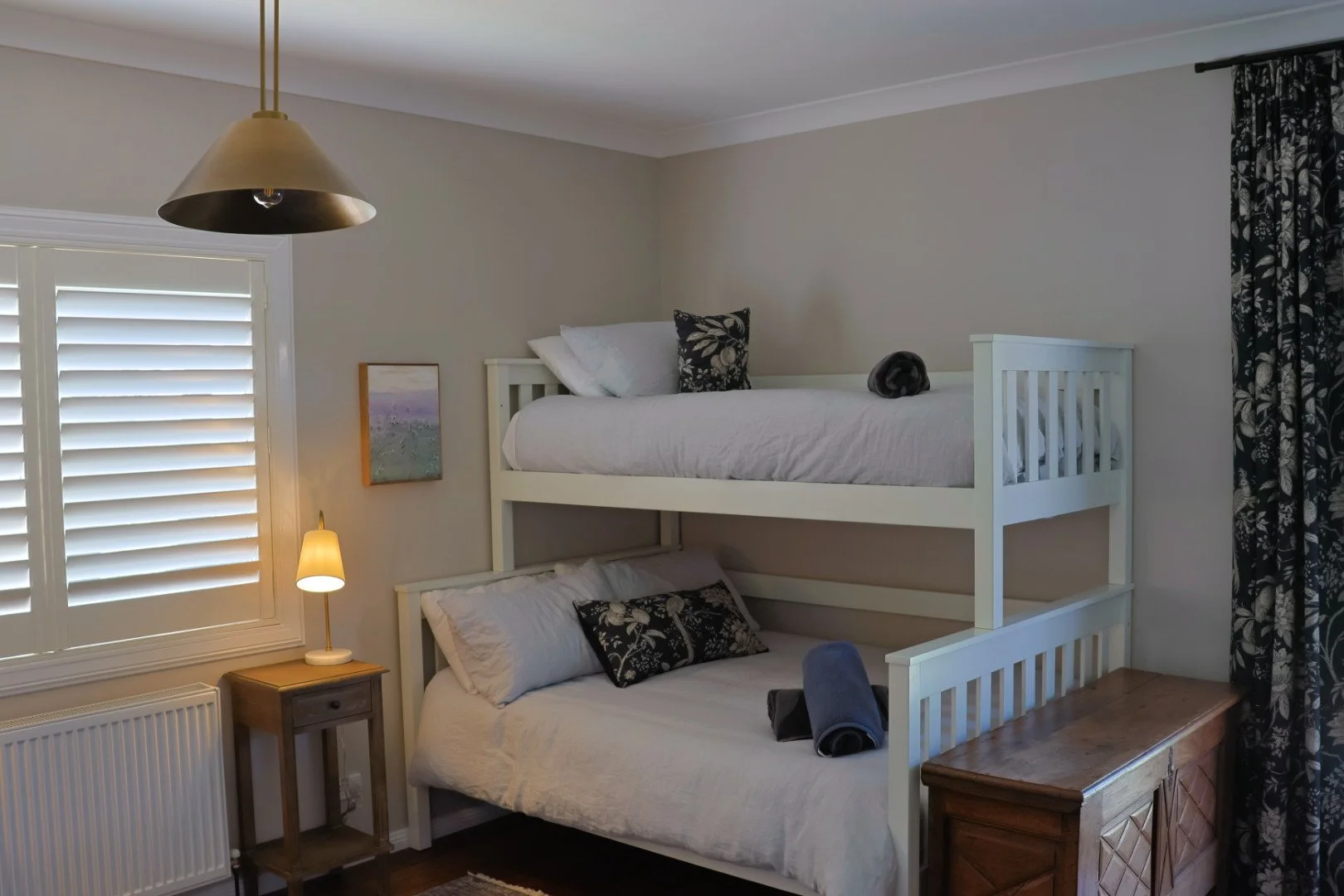 A bedroom with a bunk bed, a window with plantation shutters, a small bedside table with a lamp, and dark patterned curtains.
